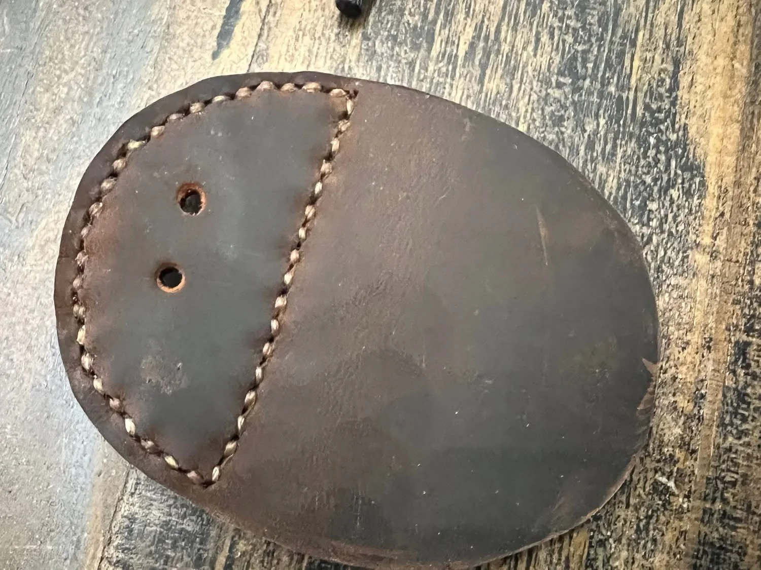 Close-up of a round, rusted metal object with two small holes and decorative stitching around an irregularly shaped area, placed on a wooden surface.