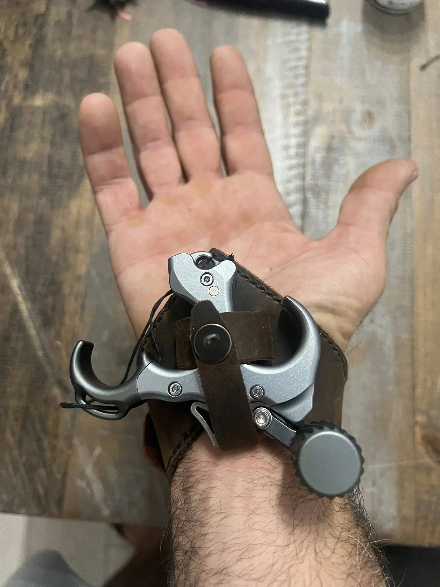 A person's hand shows a collection of climbing gear, including a karabiner, attached to a leather wrist strap, with a wooden floor and some objects in the background.