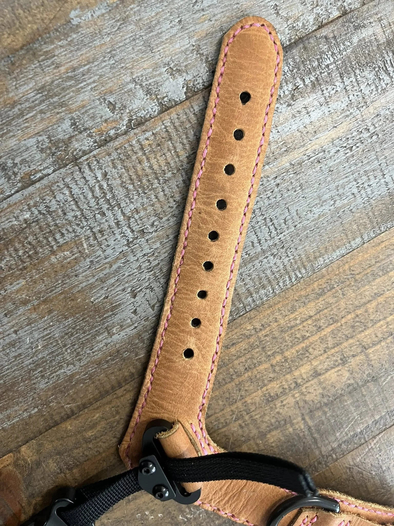 A tan leather strap with pink stitching and multiple holes, attached to a black buckle, on a wooden surface.