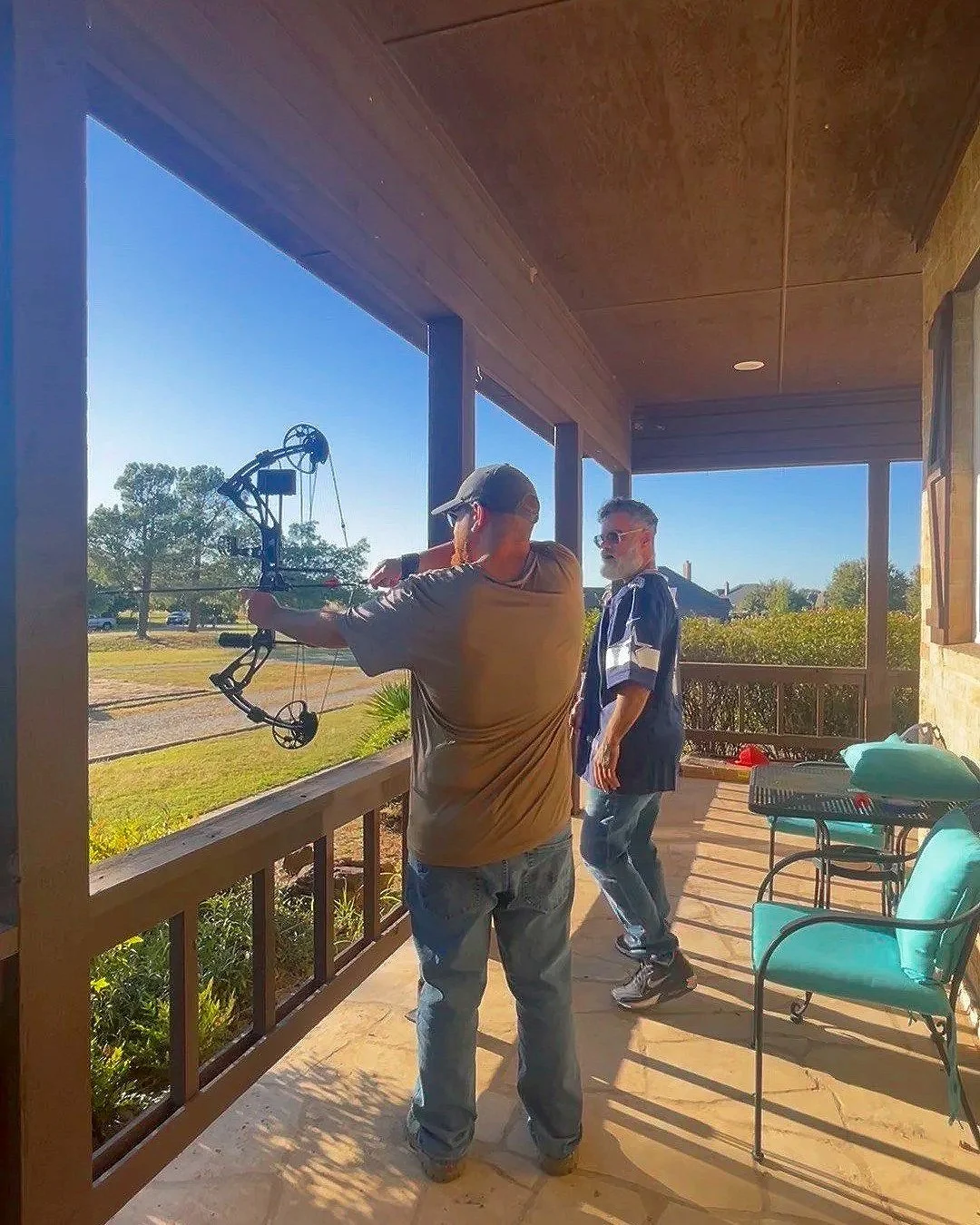 Two men practicing archery on a porch; one is drawing a bow, the other watches; outdoor scene with blue sky and trees in background.