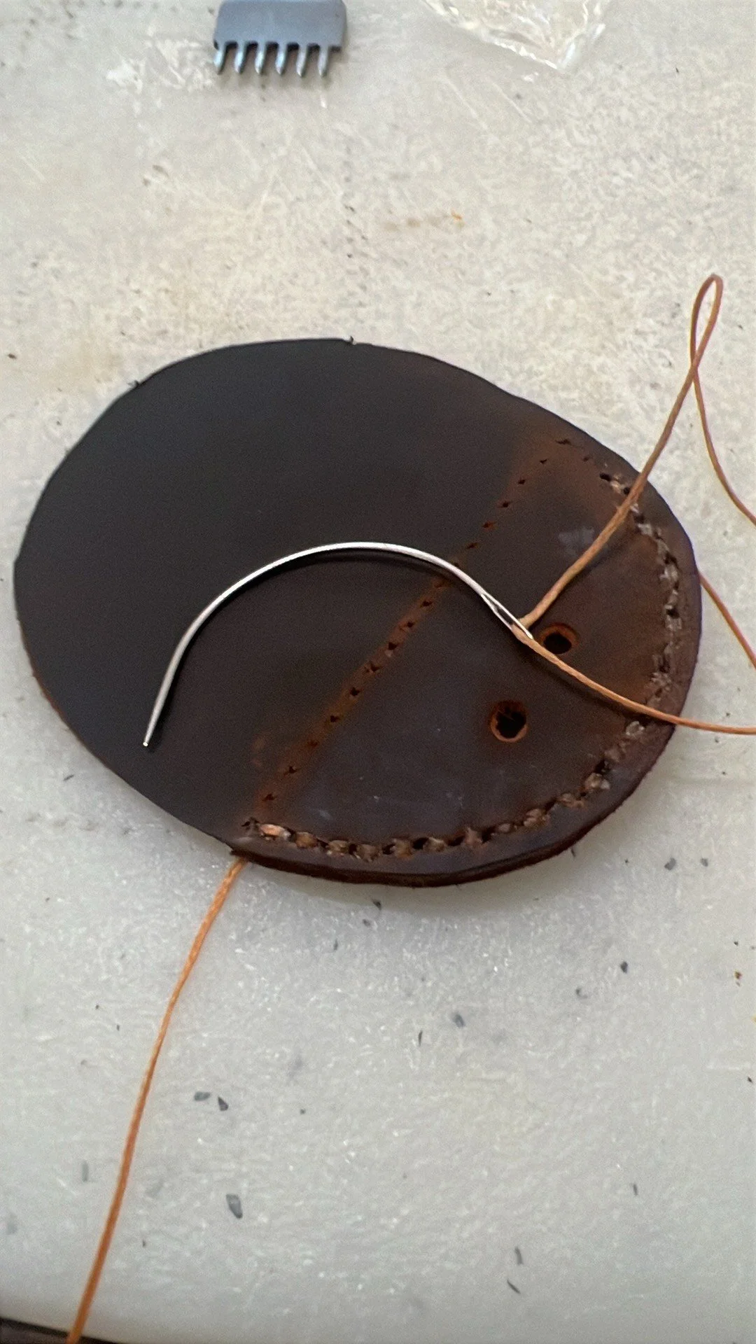 A black leather piece with stitching, a metal needle, and thin copper wire on a white surface with a small gray plastic comb in the background.