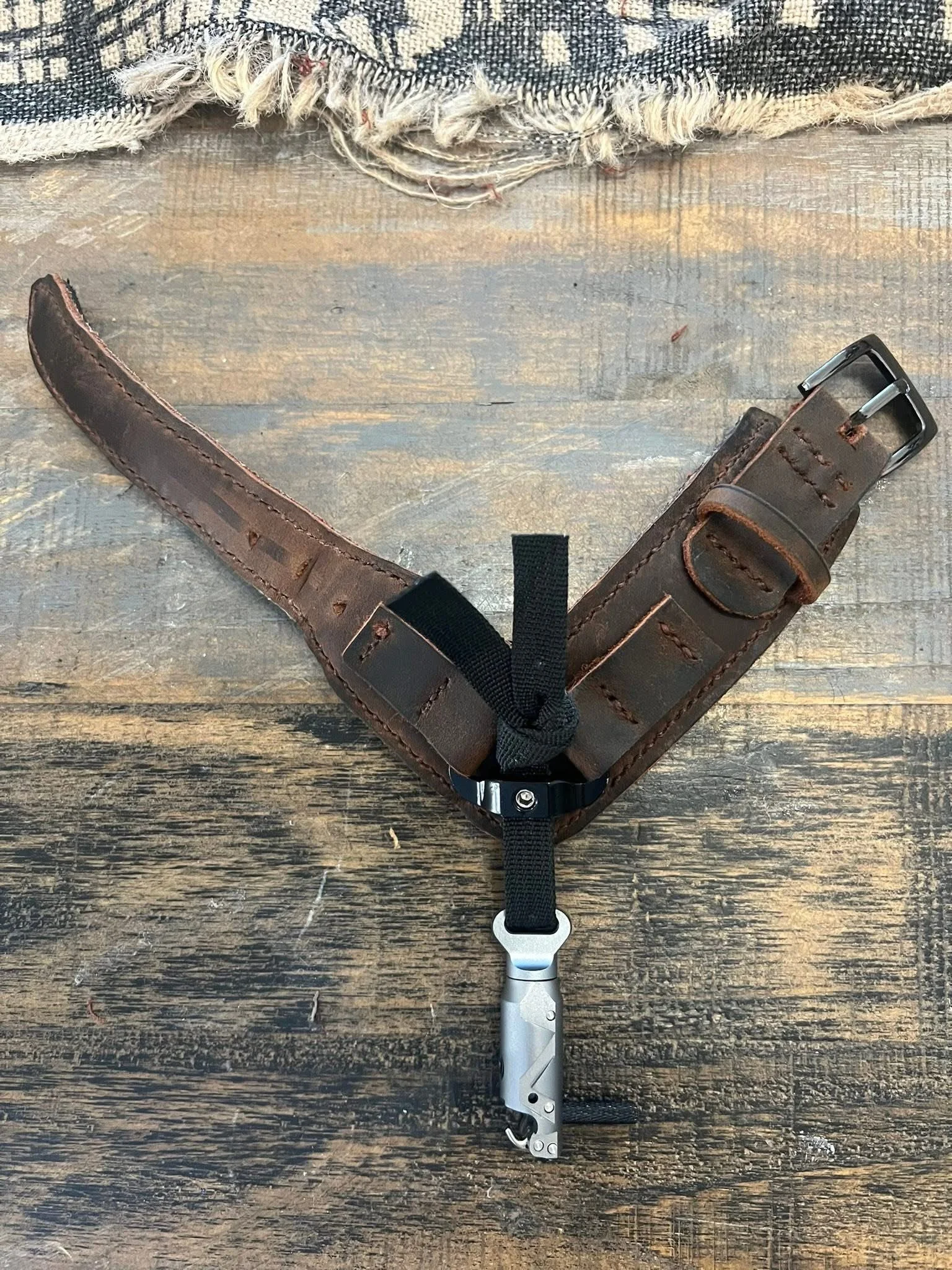 A dog harness with a metal clip, brown leather straps, and a black fabric strap on a wooden surface with a frayed textile above.