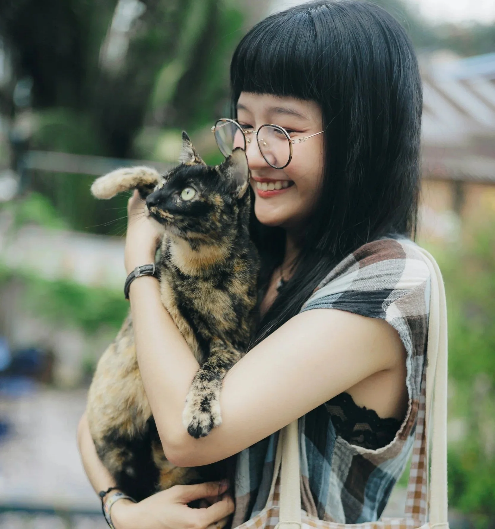 A young woman with black hair and glasses holding a tortoiseshell cat outside, smiling and looking at the cat.