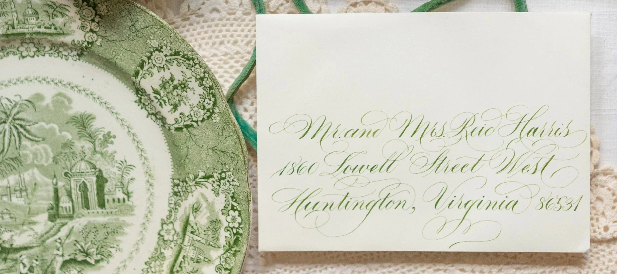 Green and white decorative plate with a scene of buildings and trees, and an elegant wedding invitation addressed to Mr. and Mrs. Reid Harris at 1860 Lowell Street West, Huntington, Virginia 86531, placed on a lace tablecloth.