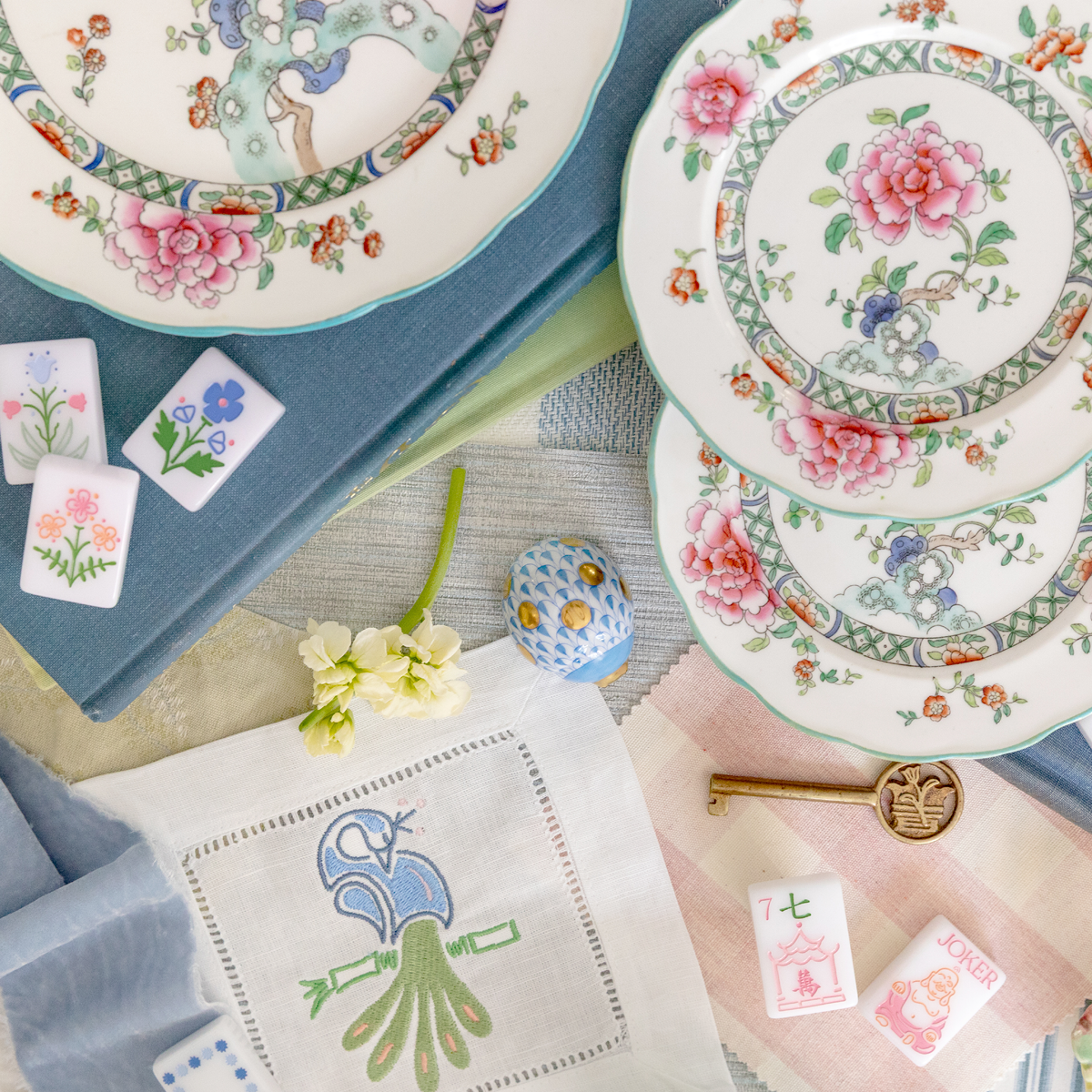 A set of decorative plates with floral and fruit patterns, a small blue and white ceramic egg, a white cloth with embroidered flower and bird design, small tiles with floral drawings, a single yellow flower, a brass key, and some playing cards with i