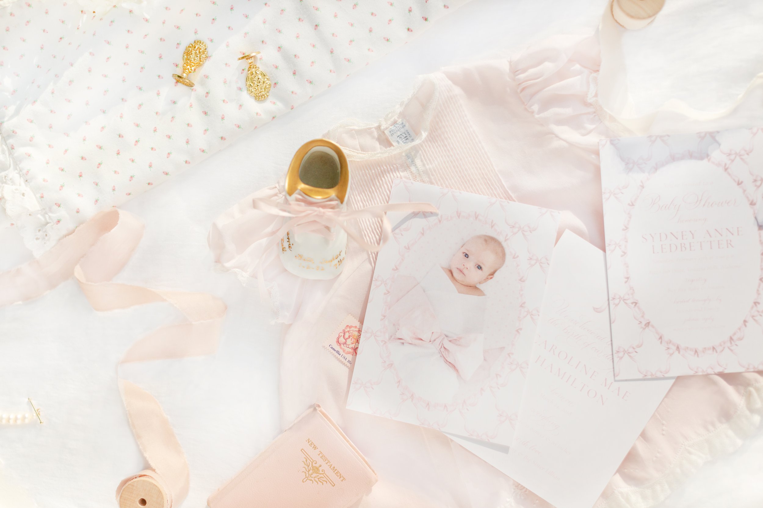 A flat lay of pink and white baby-themed items, including a baby bottle, baby cards, a ribbon, a rolled-up blanket, and gold jewelry, all arranged on a white, pink, and floral patterned background.