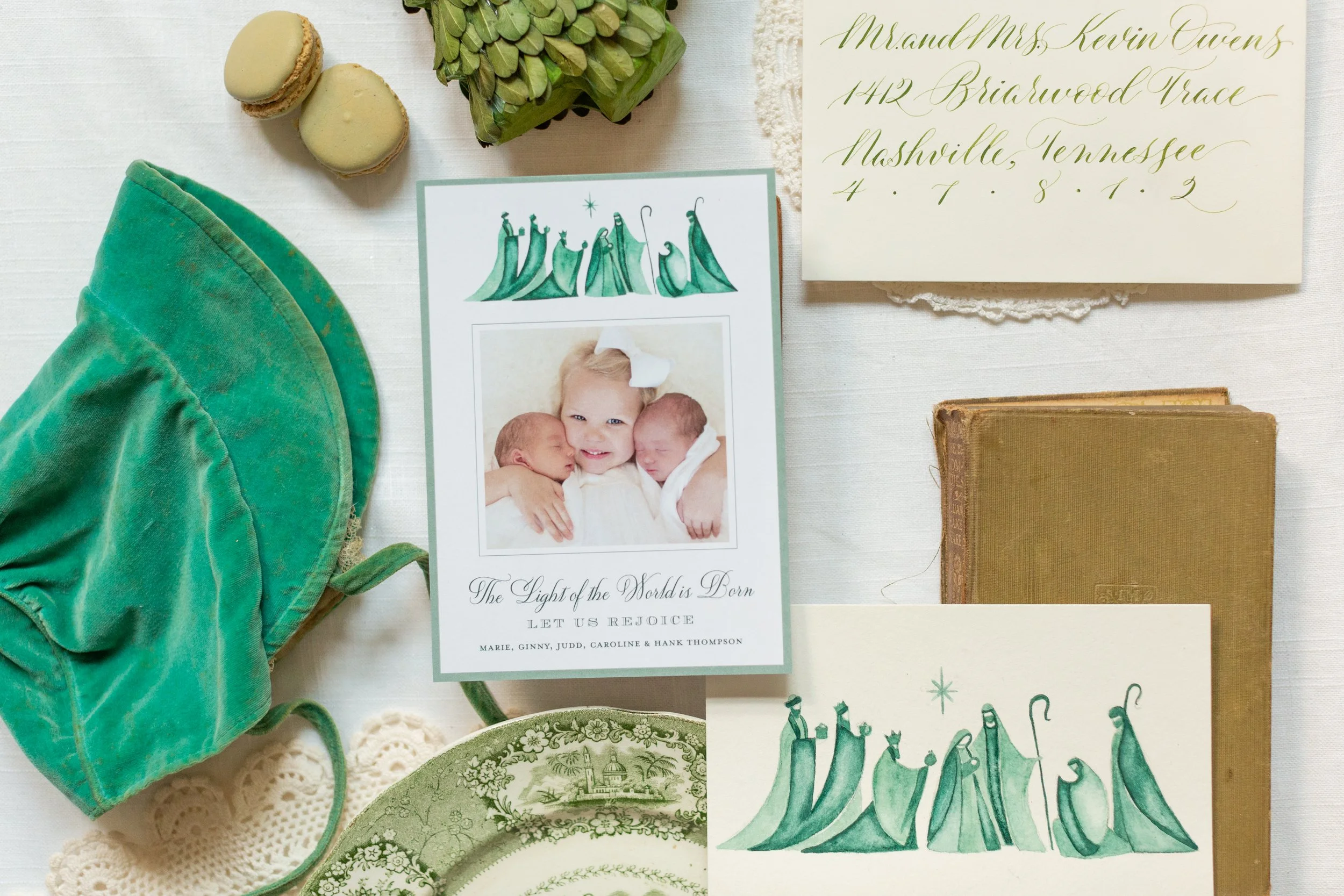 Flat lay of wedding or greeting card with green Christmas tree illustrations, a photograph of three children, and elegant gold and green calligraphy, surrounded by macarons, a succulent plant, a vintage book, and decorative dishware on a white tablecloth.
