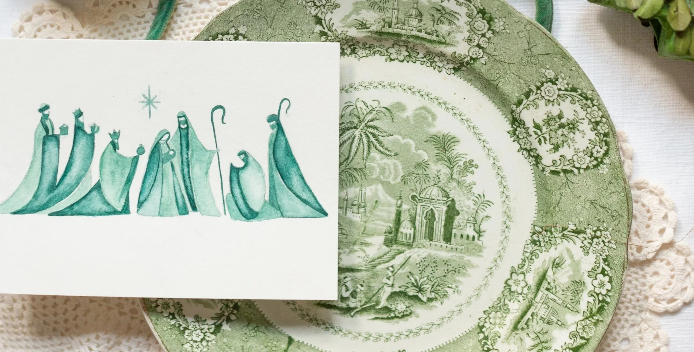 A white card with a teal watercolor illustration of the nativity scene, placed on a green decorative plate with floral and architectural patterns.