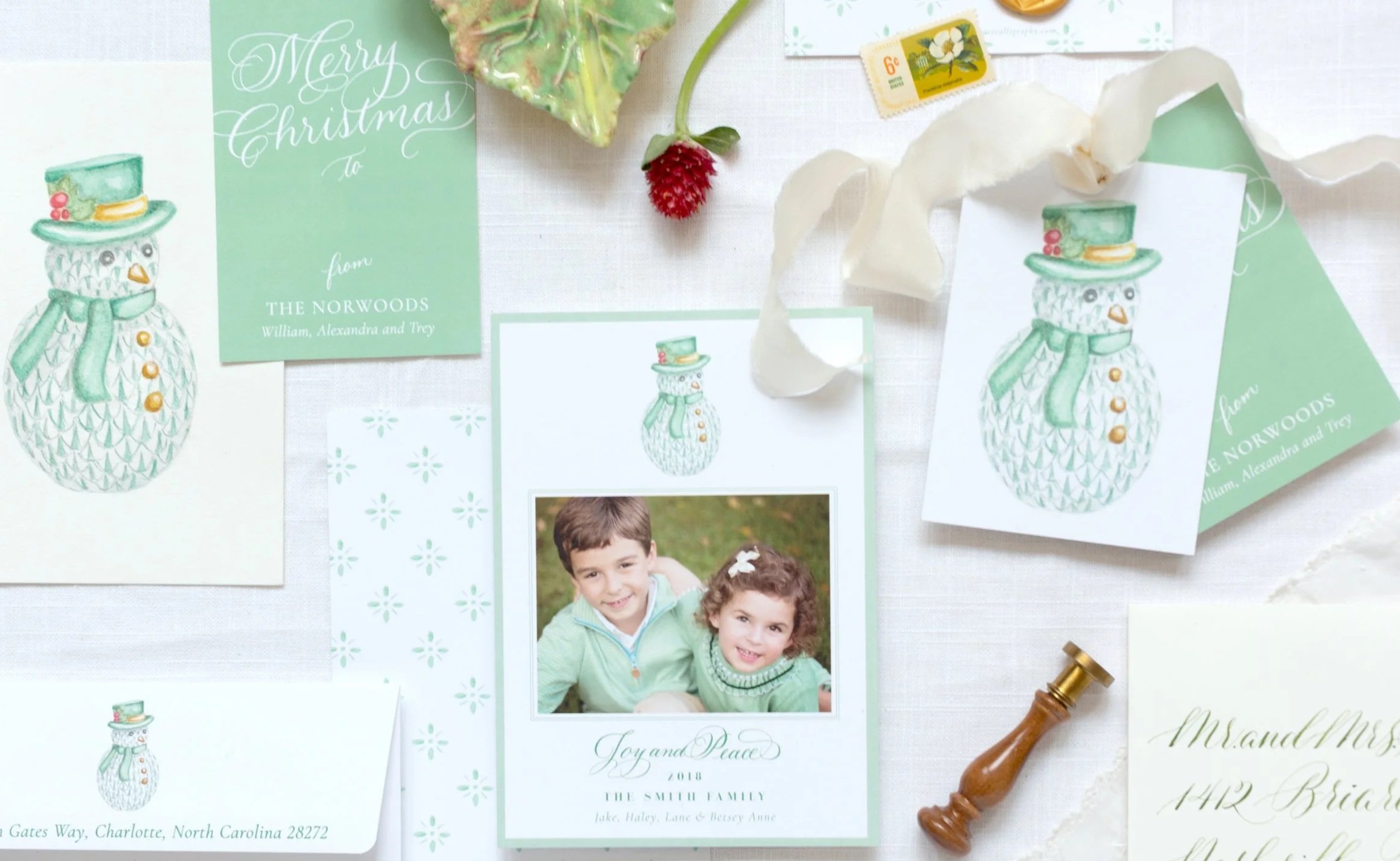 A flat lay of Christmas cards, a family photograph, a candy cane, a vanilla bean, and a small decorative snowman card with a green hat and scarf, on a white surface. The cards feature a snowman illustration, and one says 'Merry Christmas from THE NORWOODS William, Alexandra and Tey' and another says 'Joy and Peace 2018 THE SMITH FAMILY Lake, Haley, Lane & Bessey Anne'.
