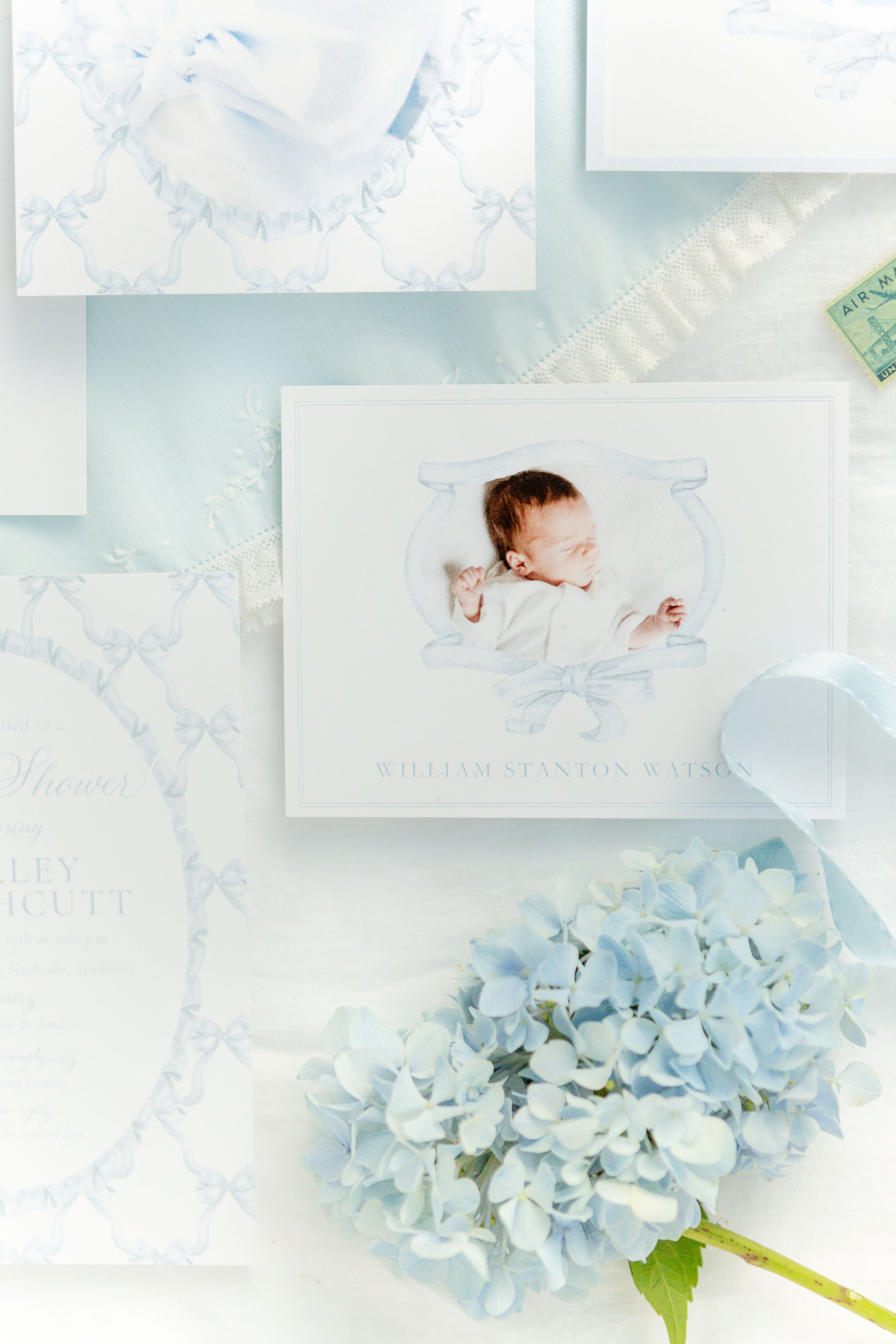 A nursery arrangement including white hydrangea flowers, a newborn photograph on a white card, and wedding stationery, all on a white lace-trimmed cloth.