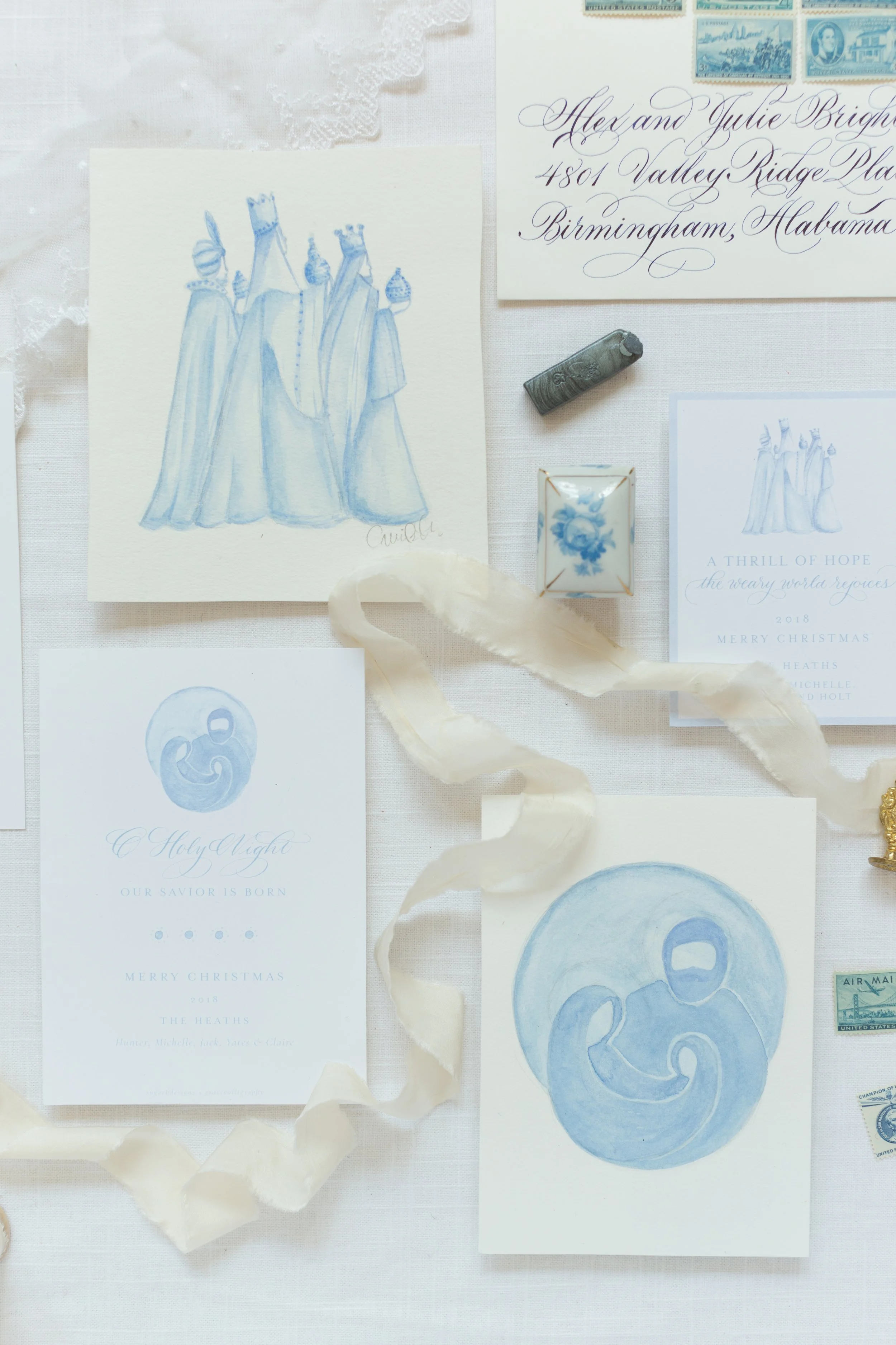 A collection of Christmas-themed greeting cards, stamps, and decorations arranged on a white fabric background, including a watercolor illustration of four women in long dresses and crowns, a calligraphy address envelope, and a ribbon.