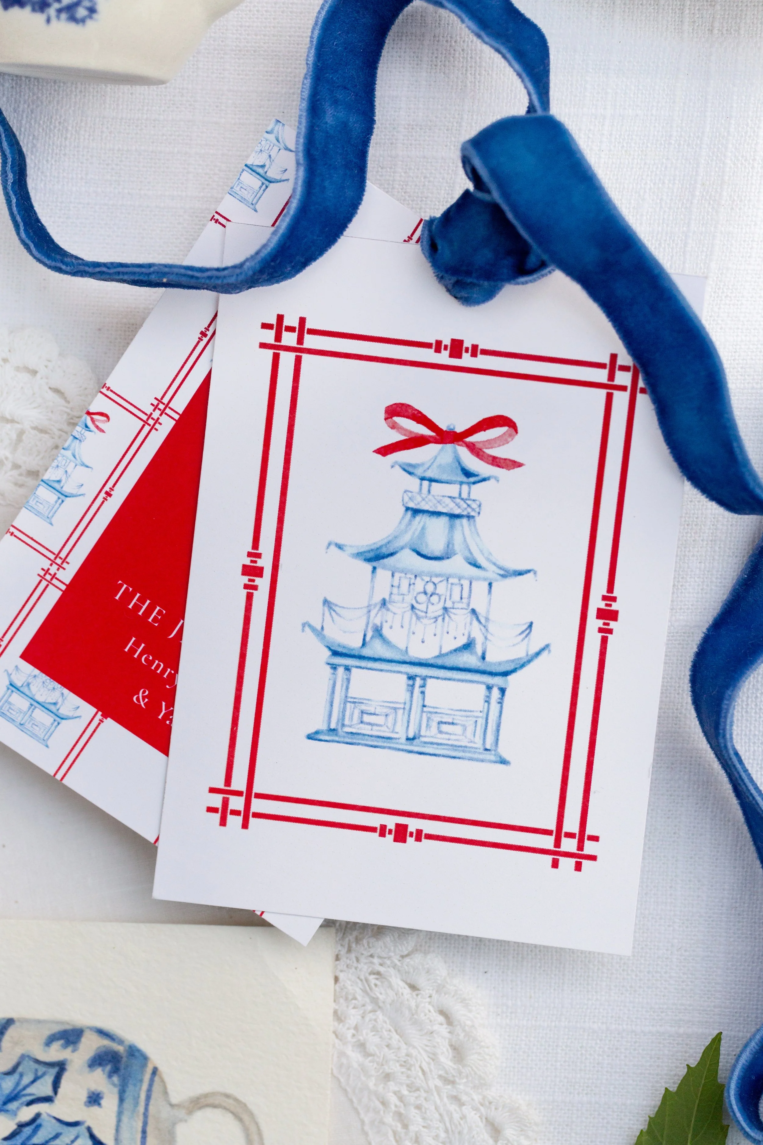 Close-up of a white card with blue and red ink illustration of a traditional Chinese lantern and pagoda, partially covered by a blue velvet ribbon, with other cards and decorative items in the background.