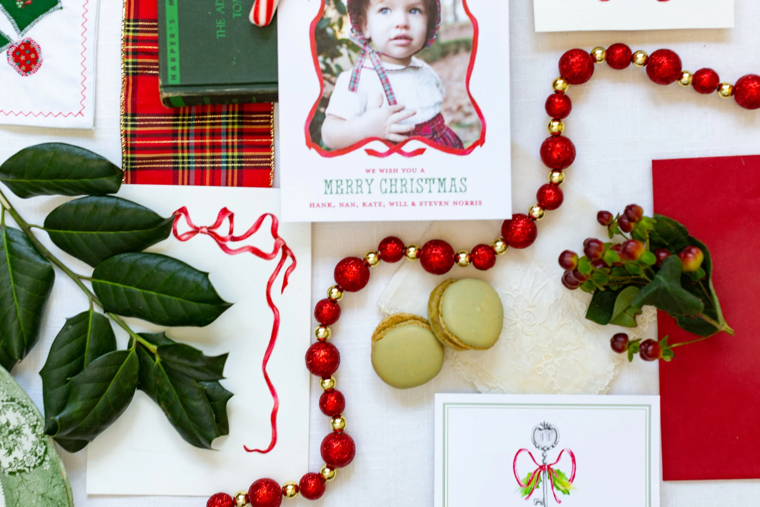 Christmas-themed items on a table including a holiday card with a child's photo, a green and red plaid ribbon, red and gold bead garland, green leaves, a red card, and cookies.
