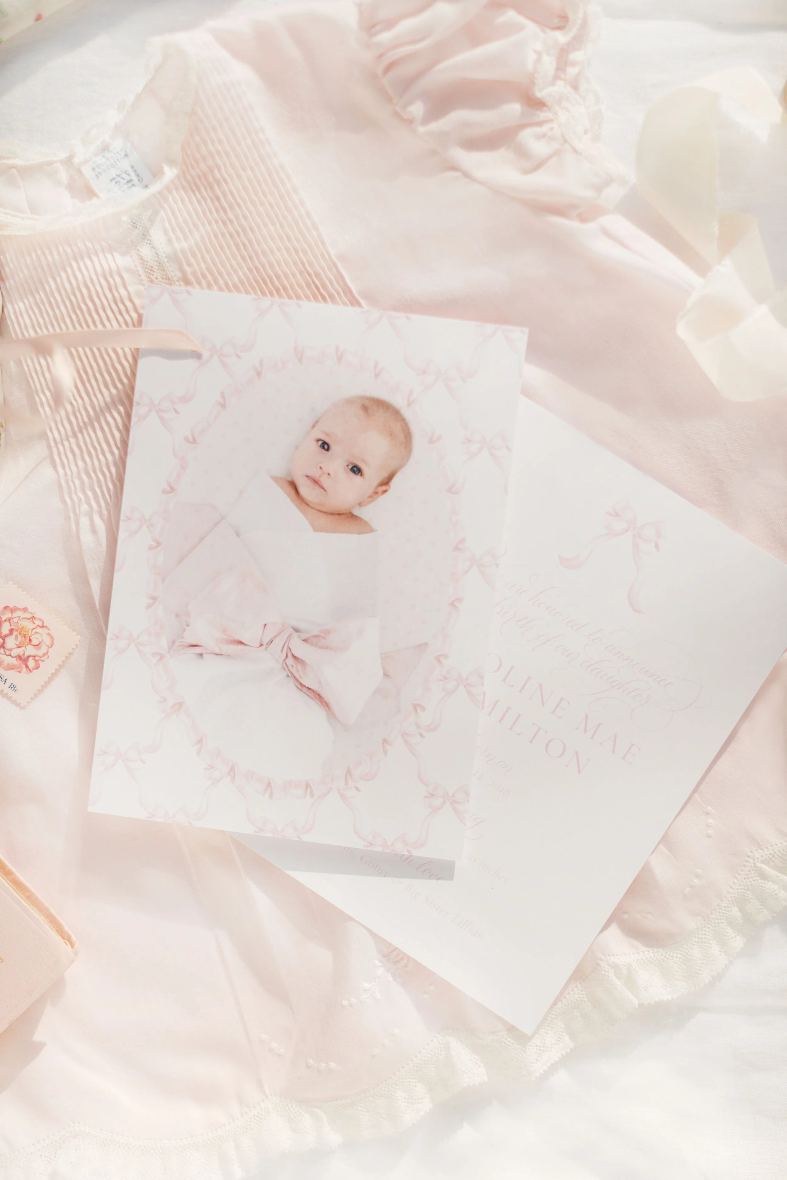 A baby portrait card with pink bows design, placed on a white satin lace fabric, accompanied by a small postage stamp.