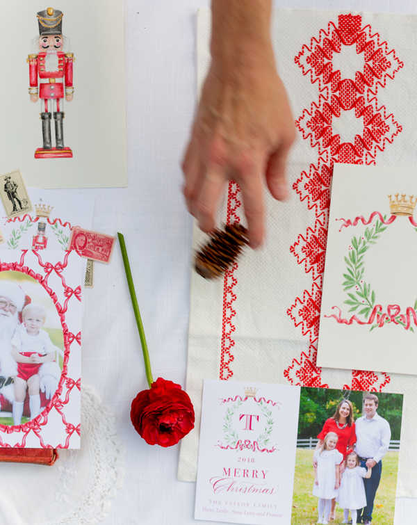 A person's hand is decorating a Christmas-themed cloth with red embroidery, surrounded by holiday cards, a red flower, and a toy soldier figurine.