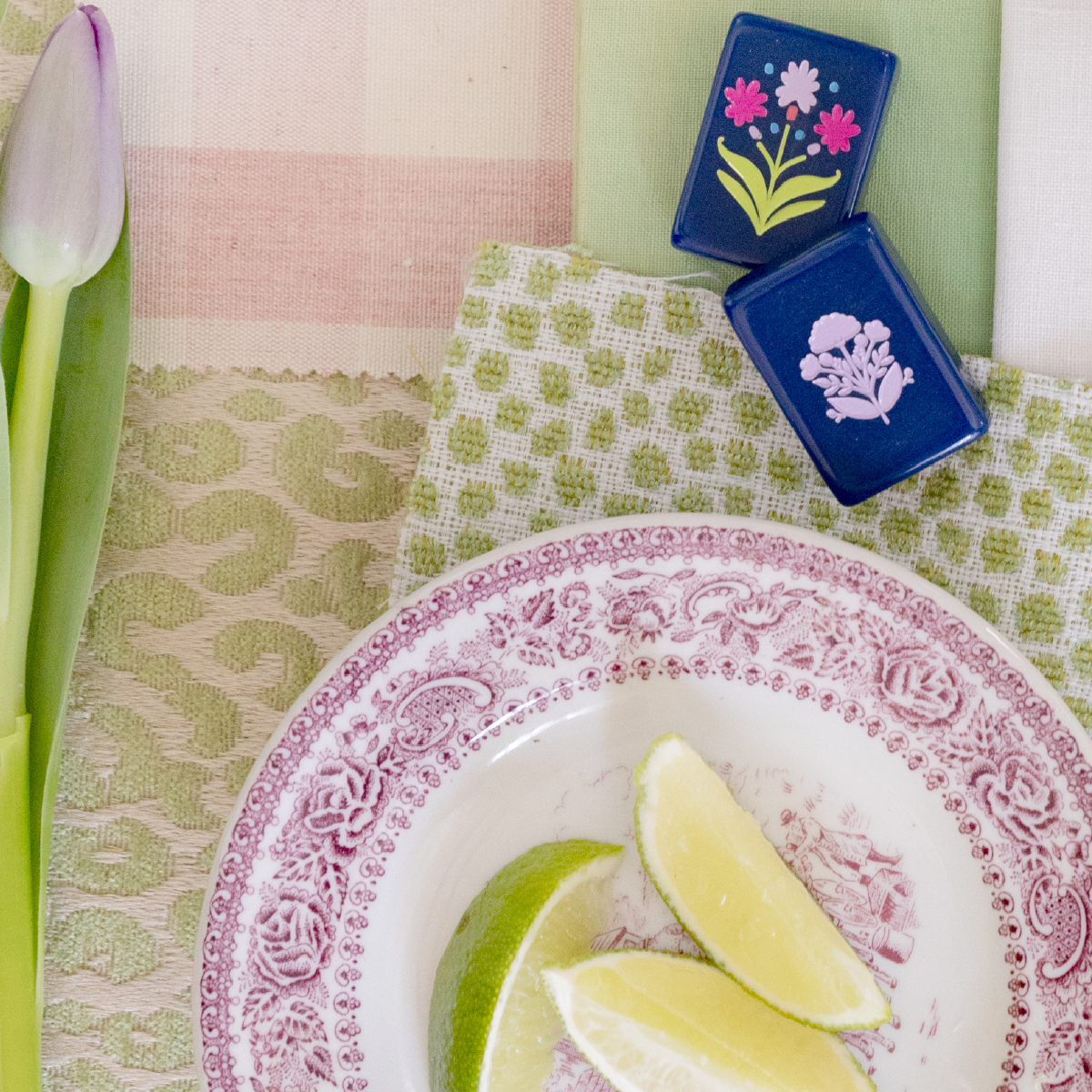 Lemon wedges on a decorative pink and white plate, with a tulip flower on the side, set on a colorful table with green and pink patterned napkins and two small blue boxes with floral designs.