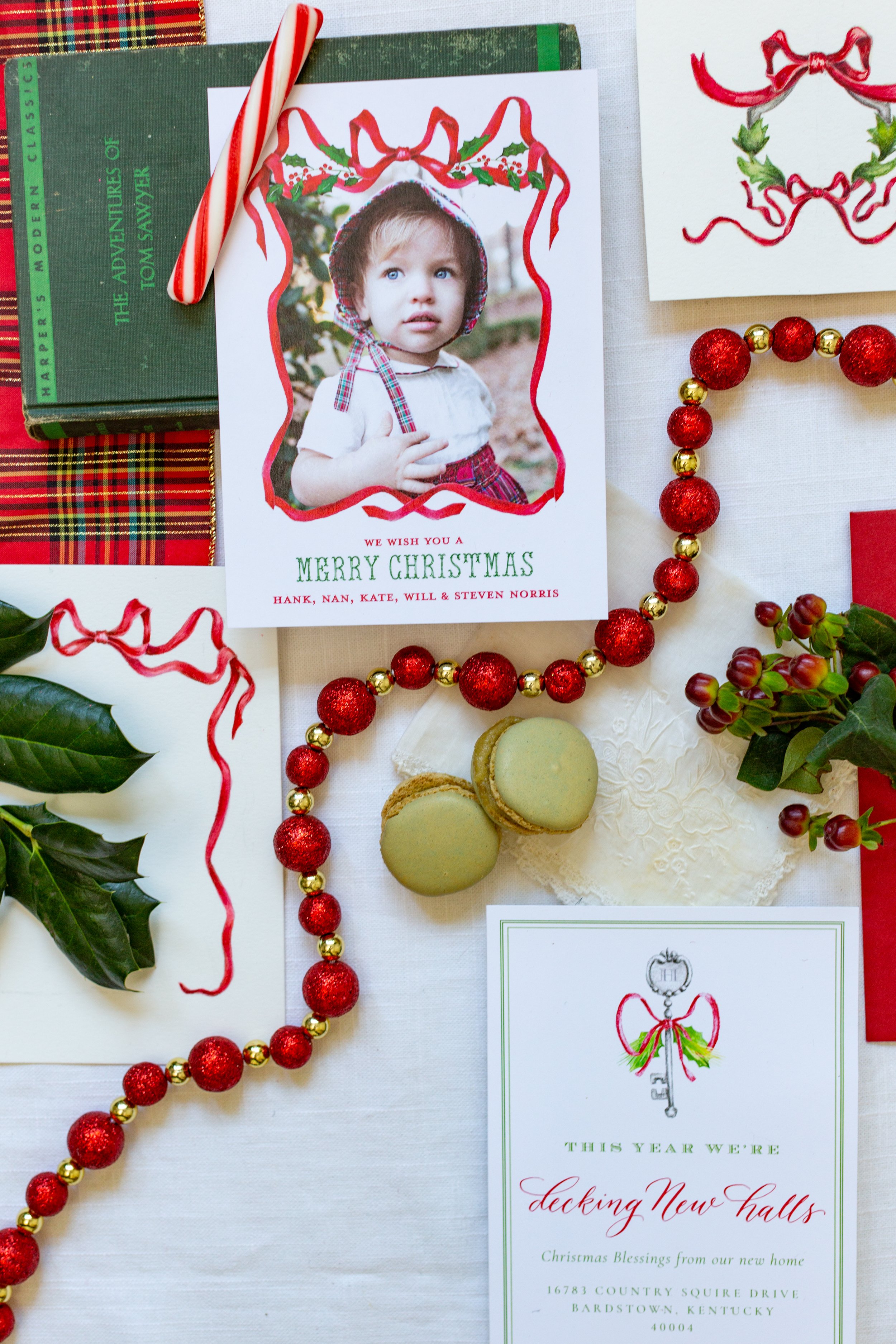 Christmas holiday card with a child's photo, holiday decorations including holly leaves, red and gold bead garland, and cookies, on a white table.