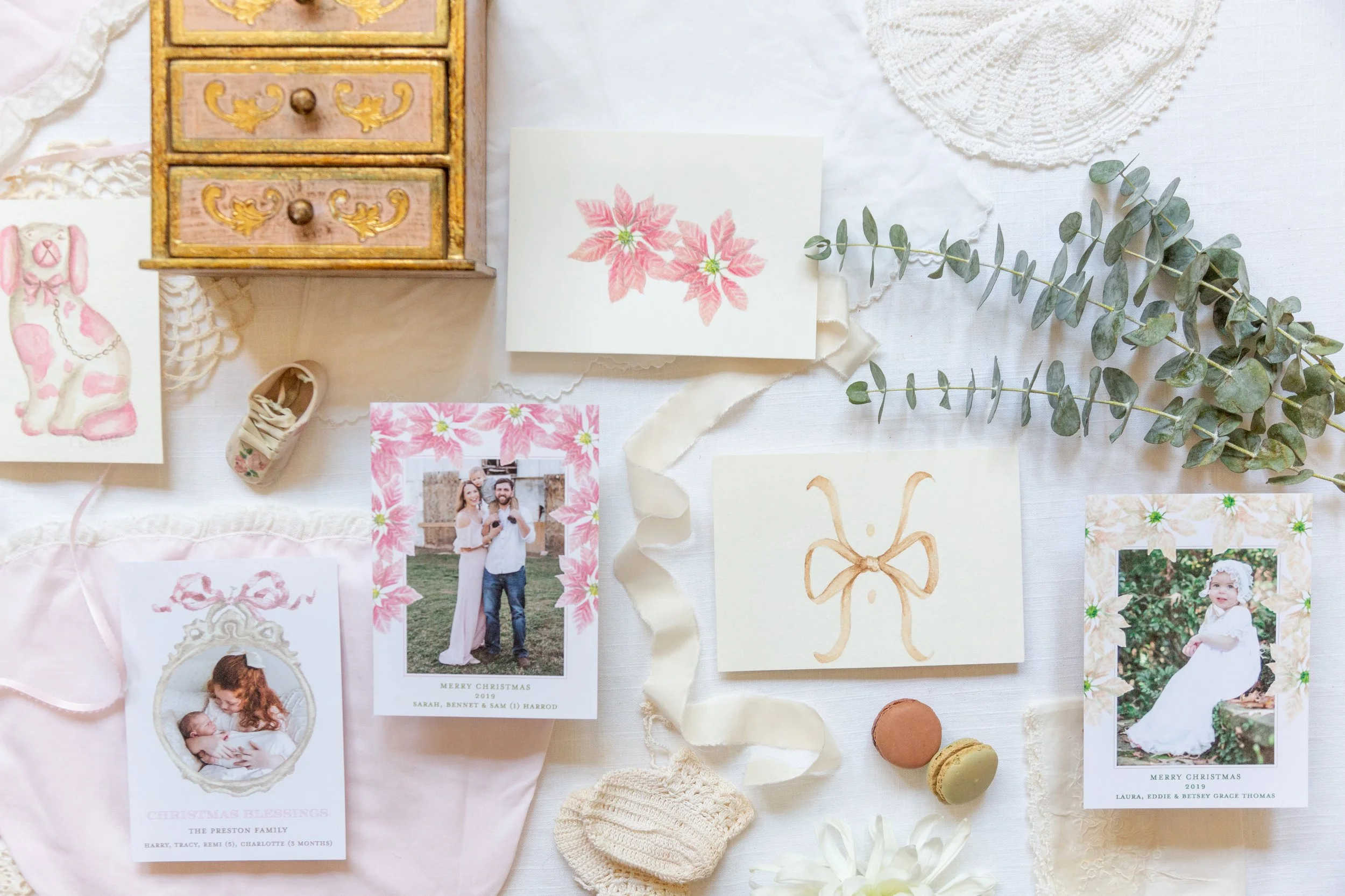 A flat lay of vintage and holiday-themed items including framed family photographs, a handmade birthday card with pink poinsettias, a crochet doily, eucalyptus sprigs, macarons, and decorative elements such as a knitted doll and a paper bunny. The images have a soft, pastel color palette.