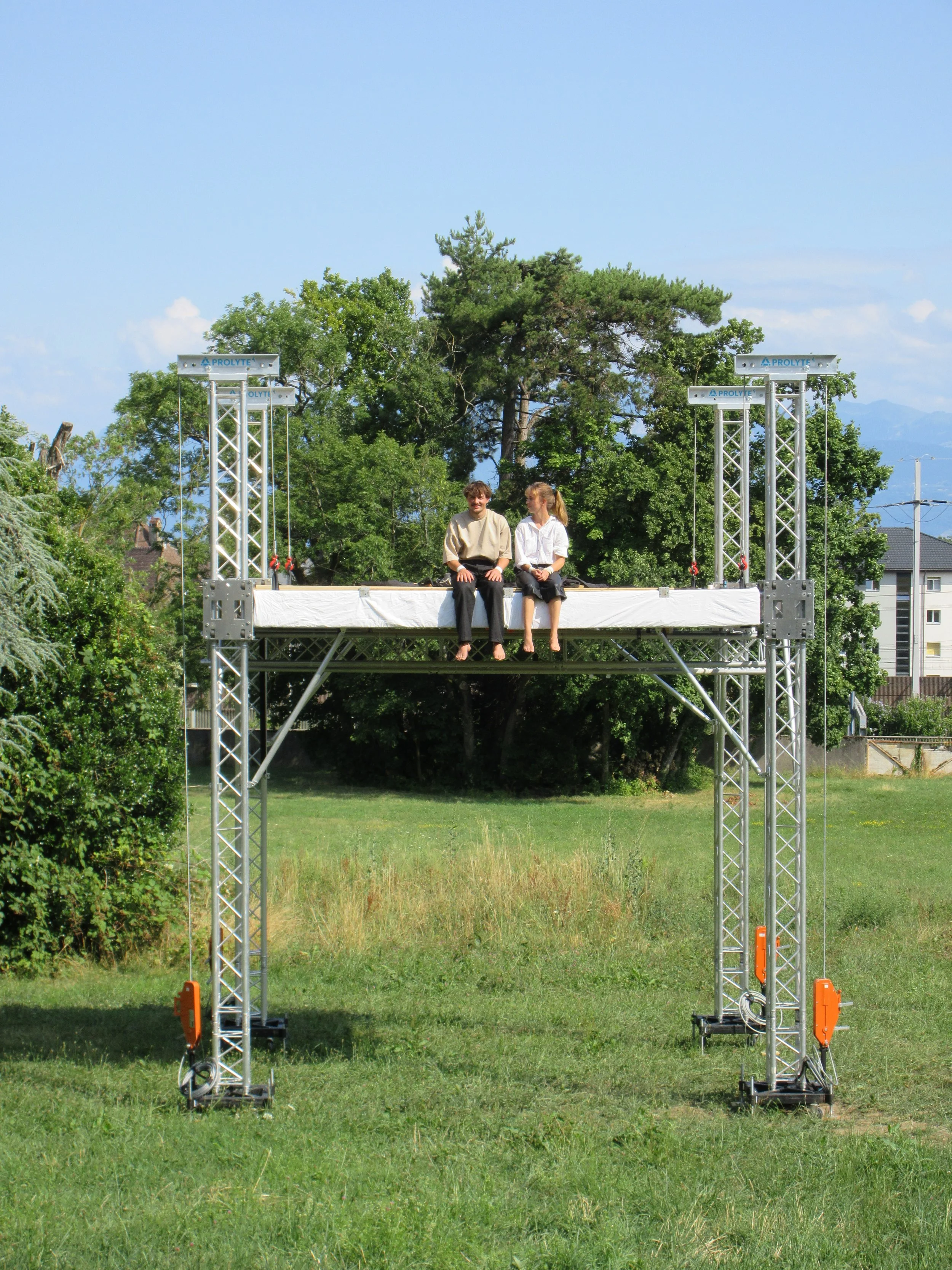 Two people sitting on a platform of a scissor lift outdoors on a grassy field with trees and houses in the background.
