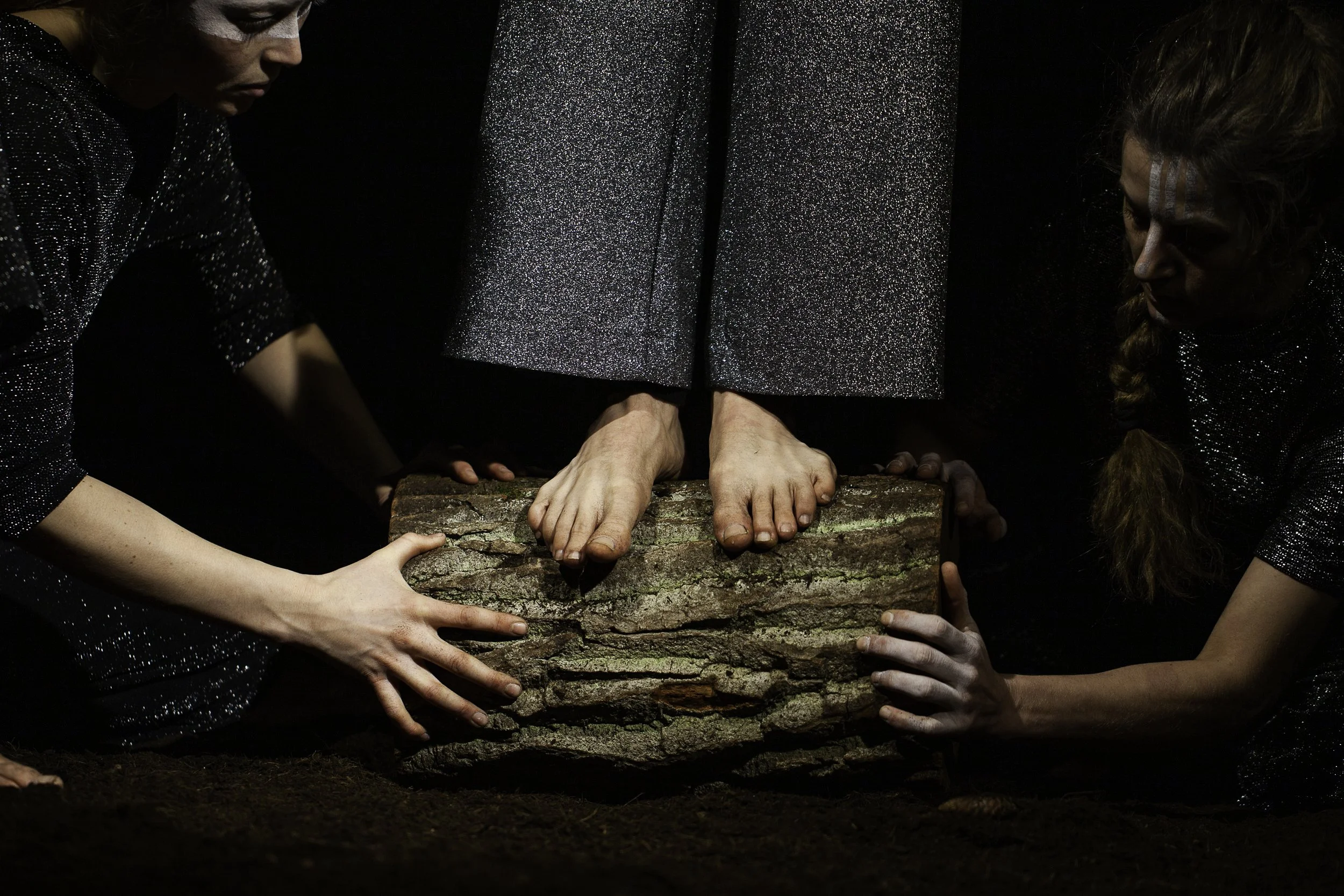 Two women wearing black, glittery outfits hold a piece of bark while another person, standing on the bark, has their feet visible, with the soles resting on the bark. The scene is dark with a black background.