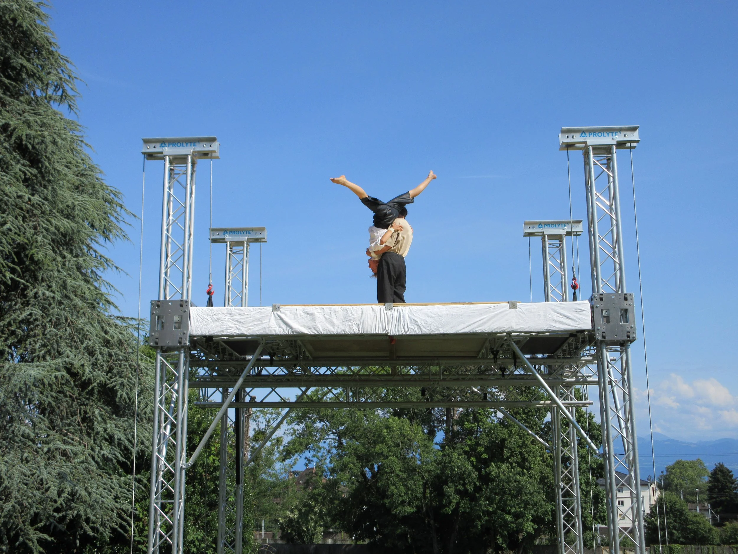 Two performers doing a handstand on a platform outdoors, with a clear blue sky and trees in the background.