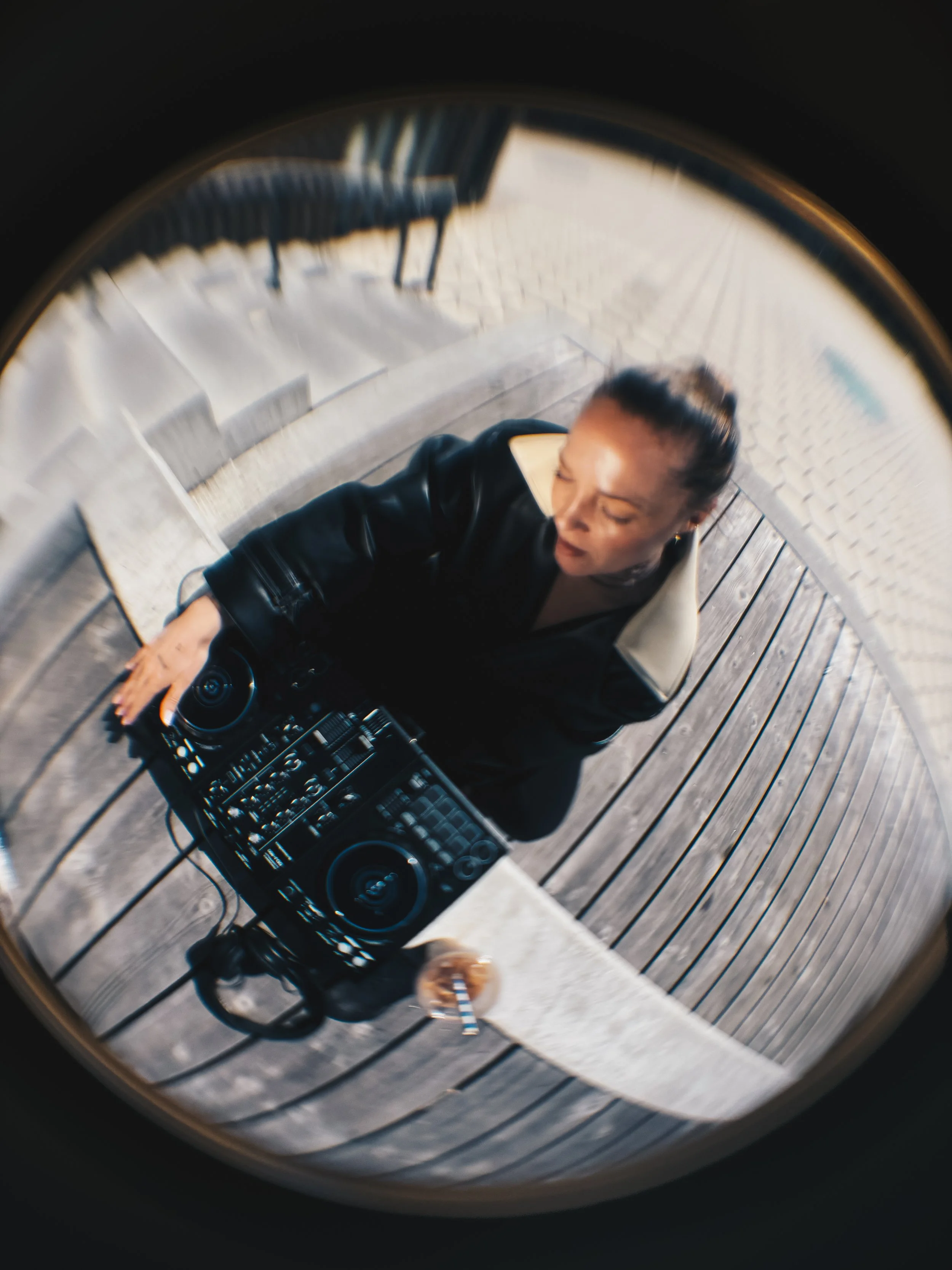A woman with dark hair pulled back, wearing a black jacket, taking a selfie with a camera or phone in a fish eye lens while working outside with her DJ equipment