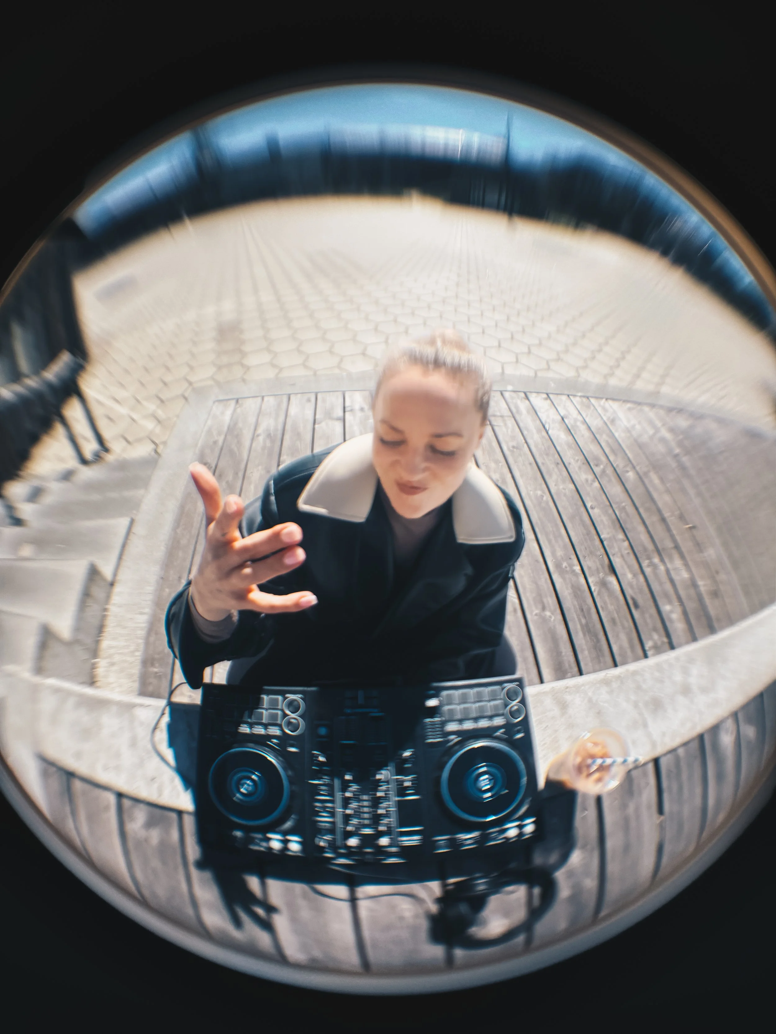 A woman with dark hair pulled back, wearing a black jacket, taking a selfie with a camera or phone in a fish eye lens while working outside with her DJ equipment