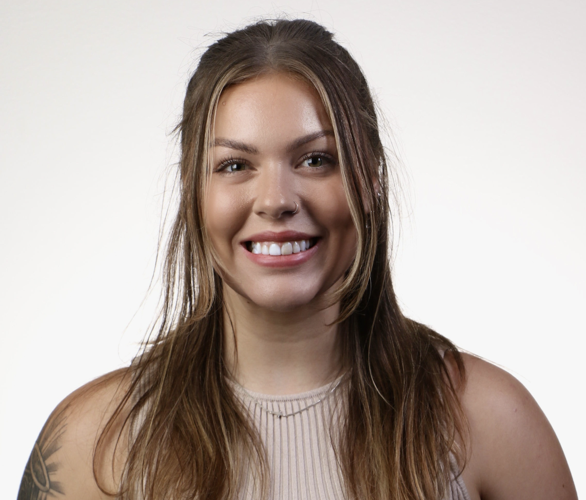 A young woman with long brown hair, smiling, wearing a sleeveless beige top and a small nose ring.
