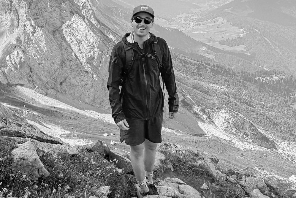 Une personne habillée en vêtements de randonnée, portant un sac à dos, un chapeau et des lunettes de soleil, se tenant sur un sentier de montagne avec un paysage accidenté et des montagnes en arrière-plan.