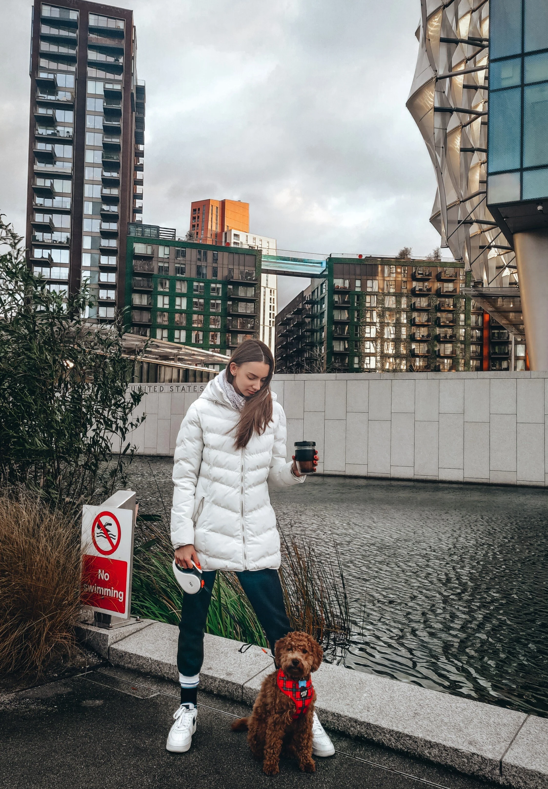 A woman in a white puffy coat holding a coffee cup and a leash with a brown curly-haired dog sitting by her, standing on a sidewalk near a body of water with modern buildings in the background. There is a 'No Swimming' sign near the water.