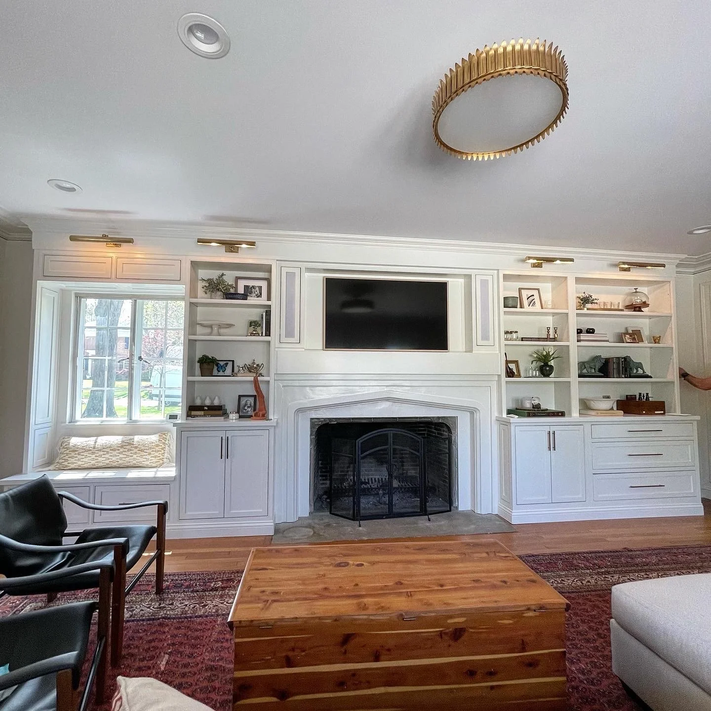 Living room with white built-in shelves around a fireplace, a flat-screen TV above the fireplace, a window seat with cushion to the left, a black leather chair to the left, a wooden coffee table in front, a white sofa to the right, a patterned rug, and a white ceiling with a gold decorative light fixture.