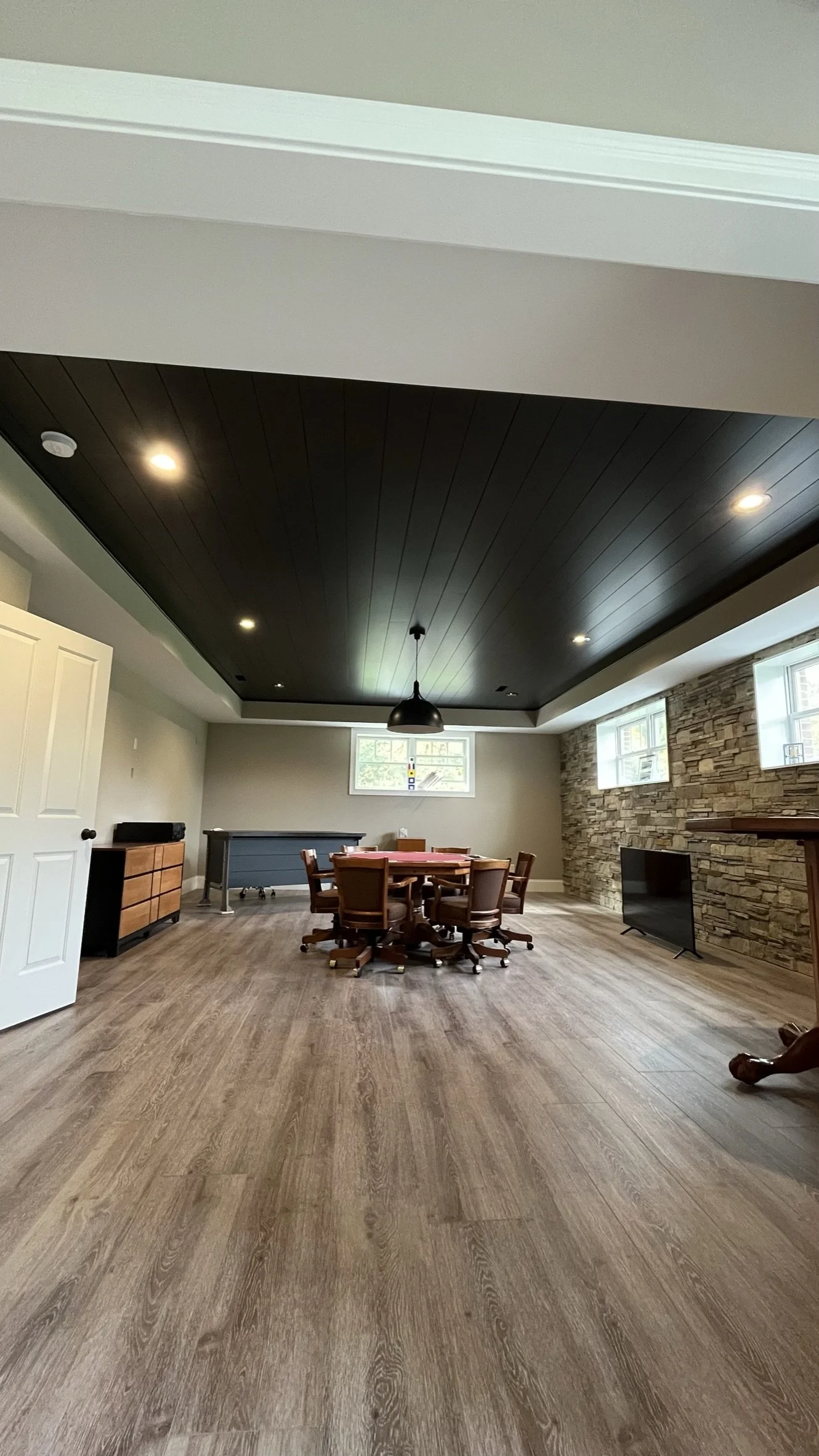 Empty game room or lounge with a round wooden table surrounded by chairs, a television on the floor, stone wall, windows, and wooden flooring.