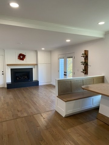 Living room with white walls, wooden flooring, a fireplace decorated with a red wreath, a set of double doors with glass panels, and a built-in bench with a wooden top.