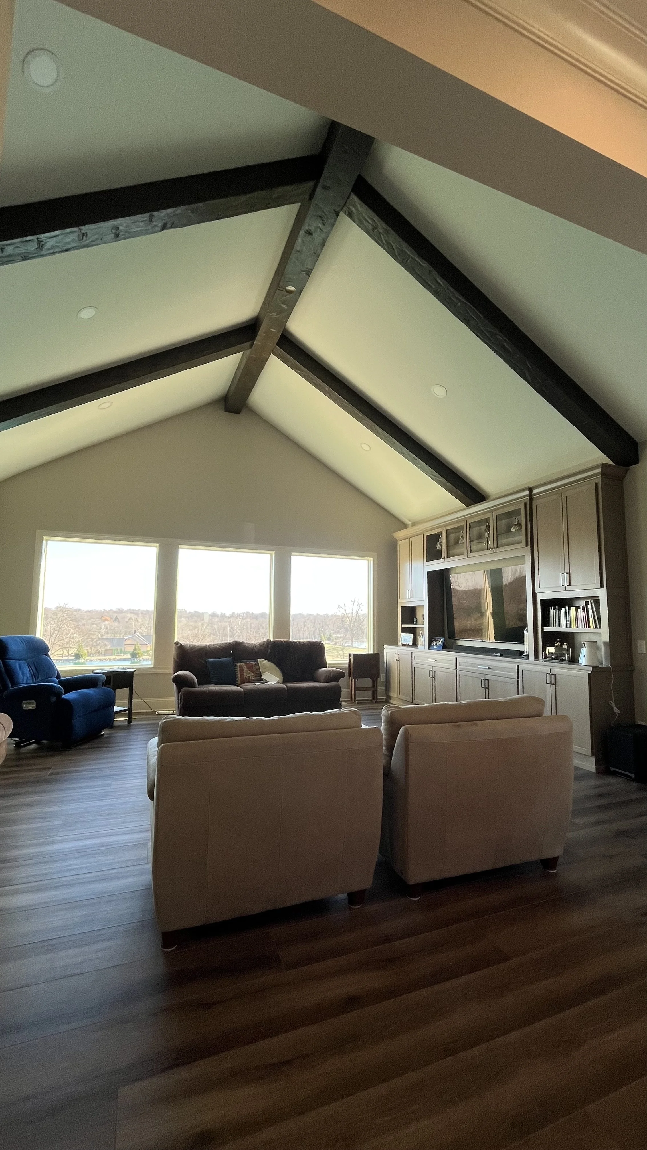 A spacious living room with a vaulted ceiling, wooden beams, a large window letting in natural light, and a beige built-in entertainment center with a TV and shelves. The room has tan upholstered chairs, a blue recliner, and a view of a landscape outside.