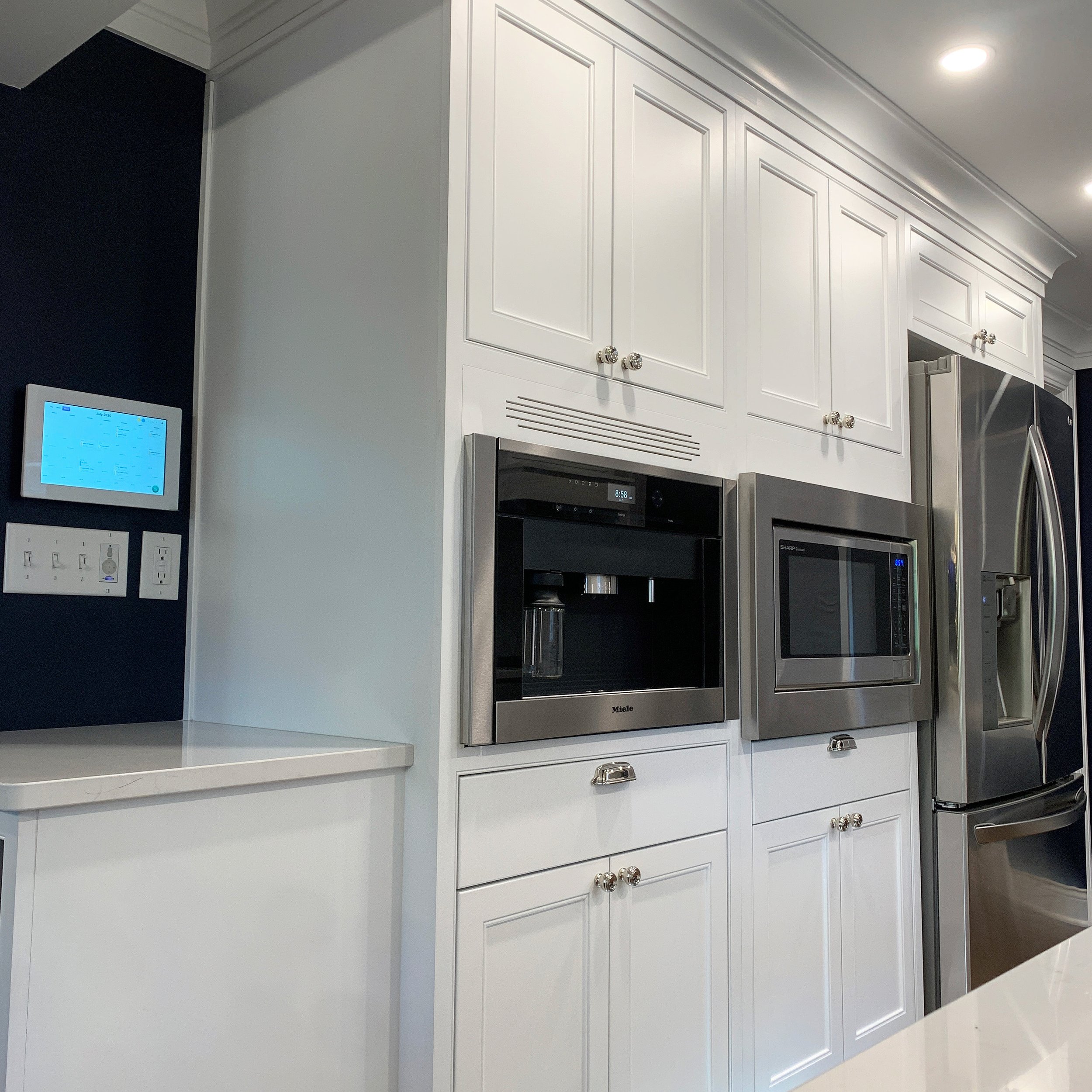 Modern kitchen with white cabinets, stainless steel microwave, coffee machine, and refrigerator, with a dark blue accent wall and digital control panel.