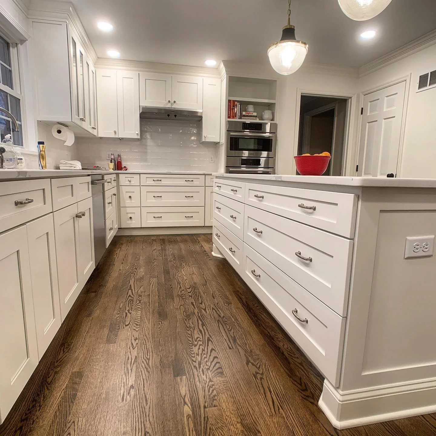 A modern kitchen with white cabinets, stainless steel appliances, and dark hardwood floors. The countertop has a red bowl, and the backsplash is made of white tiles. There is a window and ceiling lights illuminating the space.