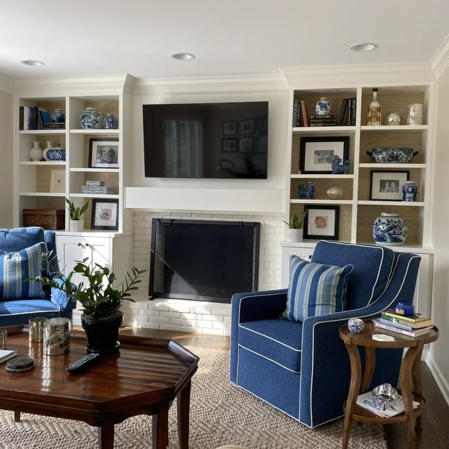 Living room with white built-in shelves, a black TV mounted above a white fireplace, blue armchairs with striped pillows, a wooden coffee table with plants and candles, and decorative items on the shelves.