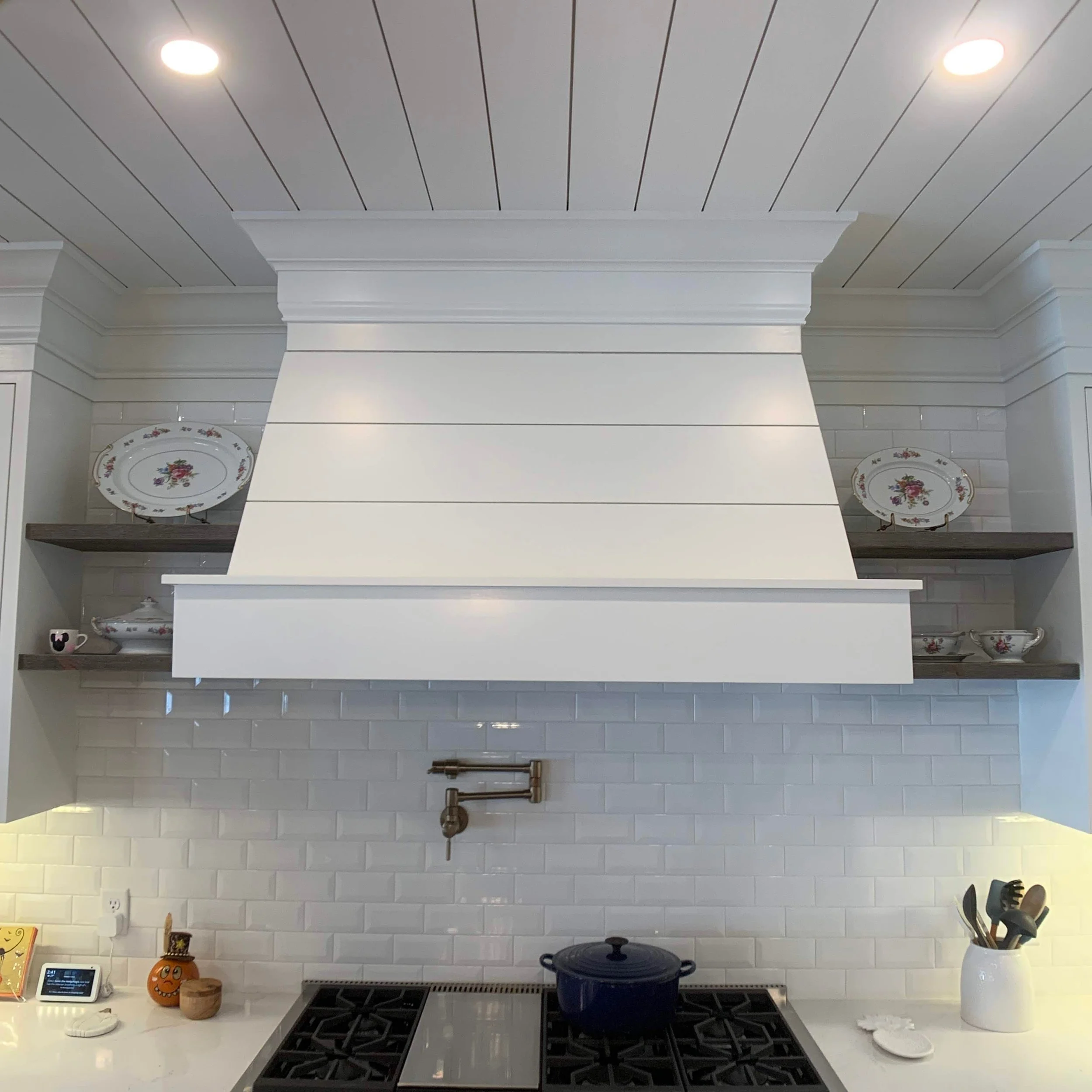 White kitchen with a stove, a white tray with decorative plates on shelves, a pot on the stove, and kitchen utensils in a white container.