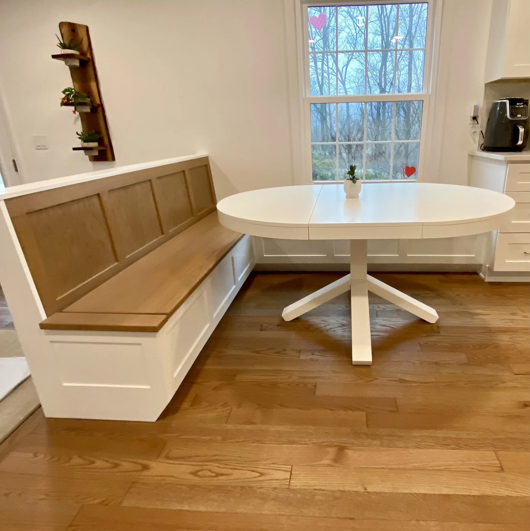 A cozy kitchen nook with a built-in wooden bench with white paneling, a white oval table with a small plant in a white pot on top, a window with trees outside, and a wooden floor.