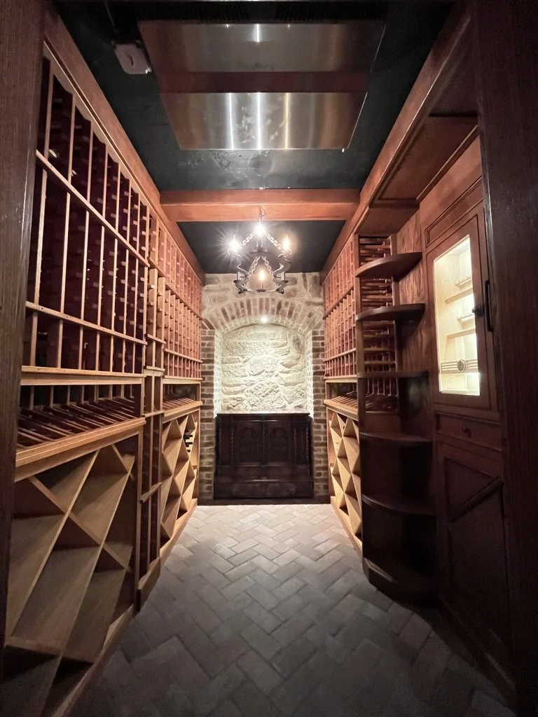 A wine cellar with wooden wine racks lining both sides, stone brick wall in the back, small window, and a wrought iron chandelier hanging from the ceiling.