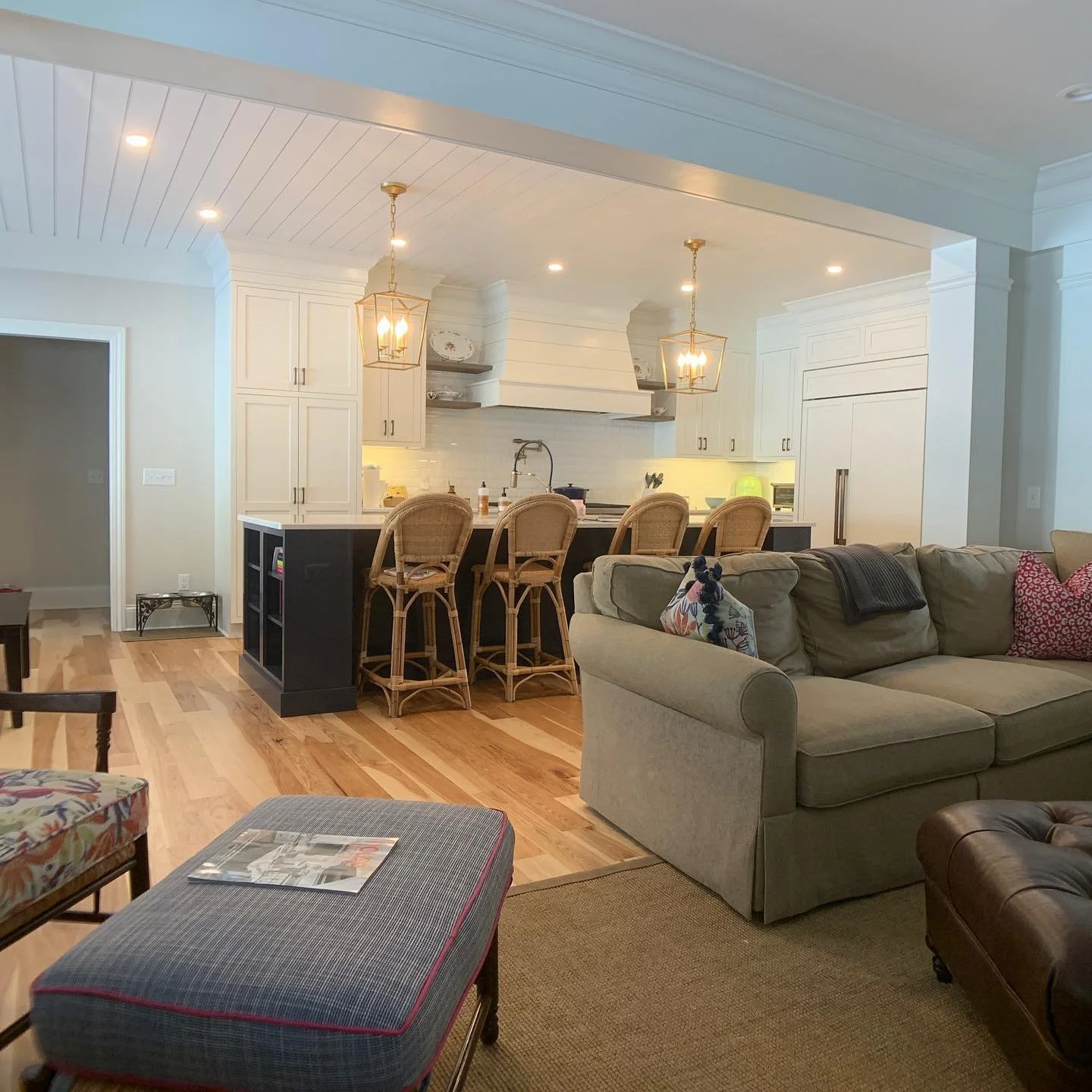 Open-concept living room and kitchen with hardwood floors, beige sofa with colorful pillows, dark wood coffee table with magazines, kitchen island with four wicker chairs, white cabinets, and pendant lighting.