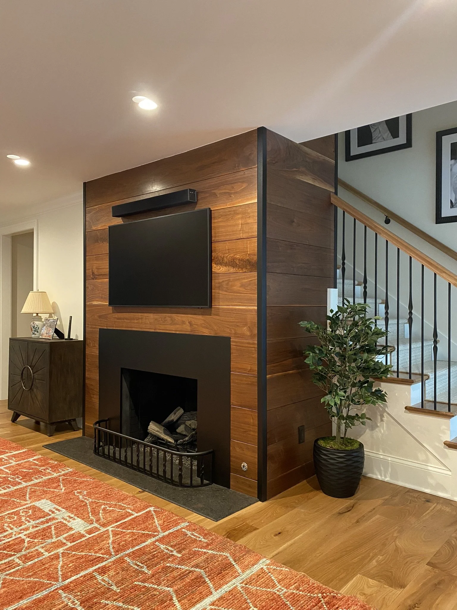 Living room with wood-paneled wall, mounted TV, fireplace with black frame, orange patterned rug, potted plant, staircase with wooden handrail and black balusters, and a console table with framed photos and a lamp in the background.
