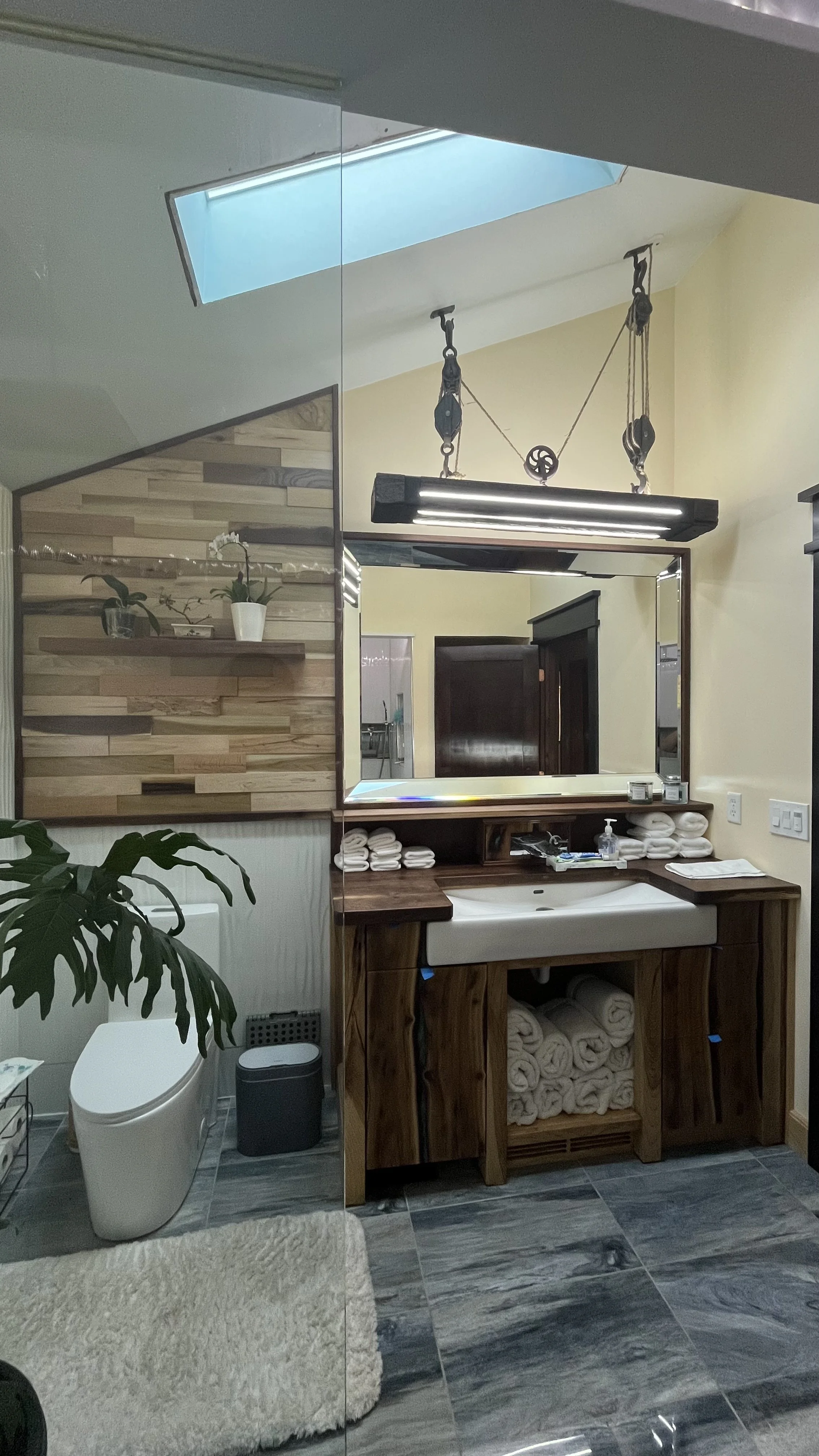 Modern bathroom with a wooden vanity, a large mirror, folded towels, a sink, and a skylight. There are plants and a white toilet with a fluffy rug in front.