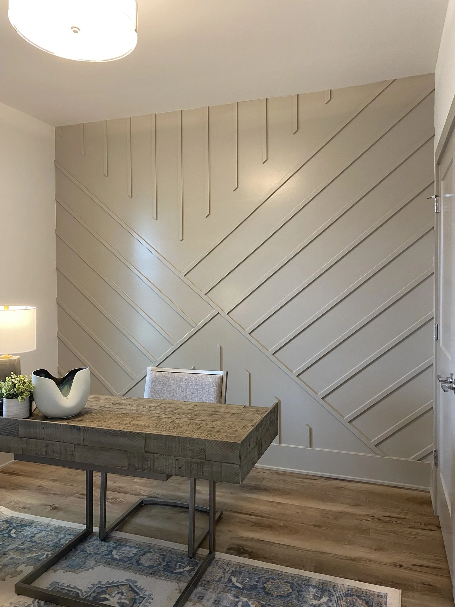 A modern interior space featuring a neutral-colored wall with geometric wooden paneling, a wooden desk with metal legs, a beige upholstered chair, a table lamp, a small plant, and decorative items on the desk, with a blue and beige patterned rug on a wooden floor.