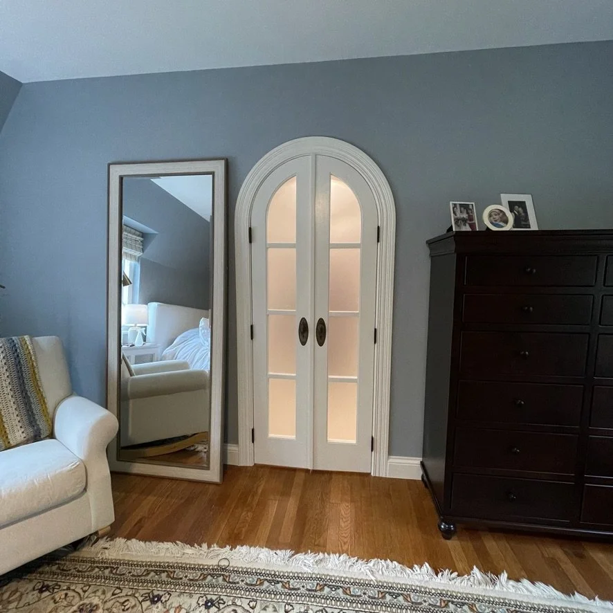 A bedroom corner with a large mirror, a white armchair reflected in the mirror, a black dresser with framed photos on top, an arched double door with light shining through, and a patterned area rug on wooden flooring.