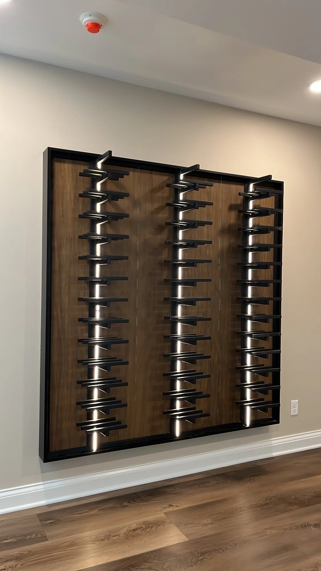 Wall-mounted bottle rack with multiple tiers of black shelves and LED lighting in a room with wood flooring and light-colored walls.