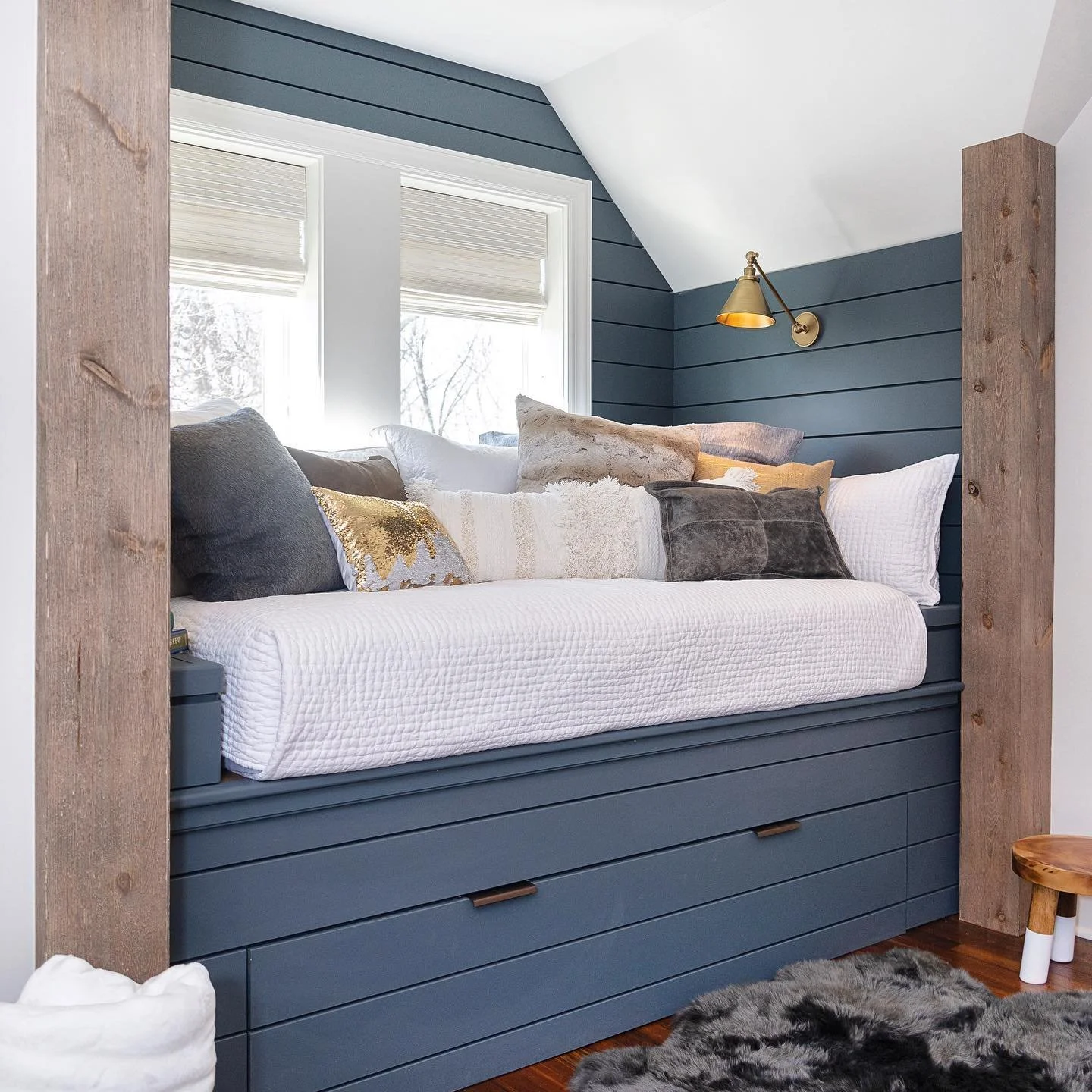 A cozy window seat with a white quilted cushion, multiple pillows in various shades including gray, gold, and beige, built into a nook with blue shiplap walls, a brass wall light, two windows with white blinds, a small wooden stool, and a gray shaggy rug on a hardwood floor.