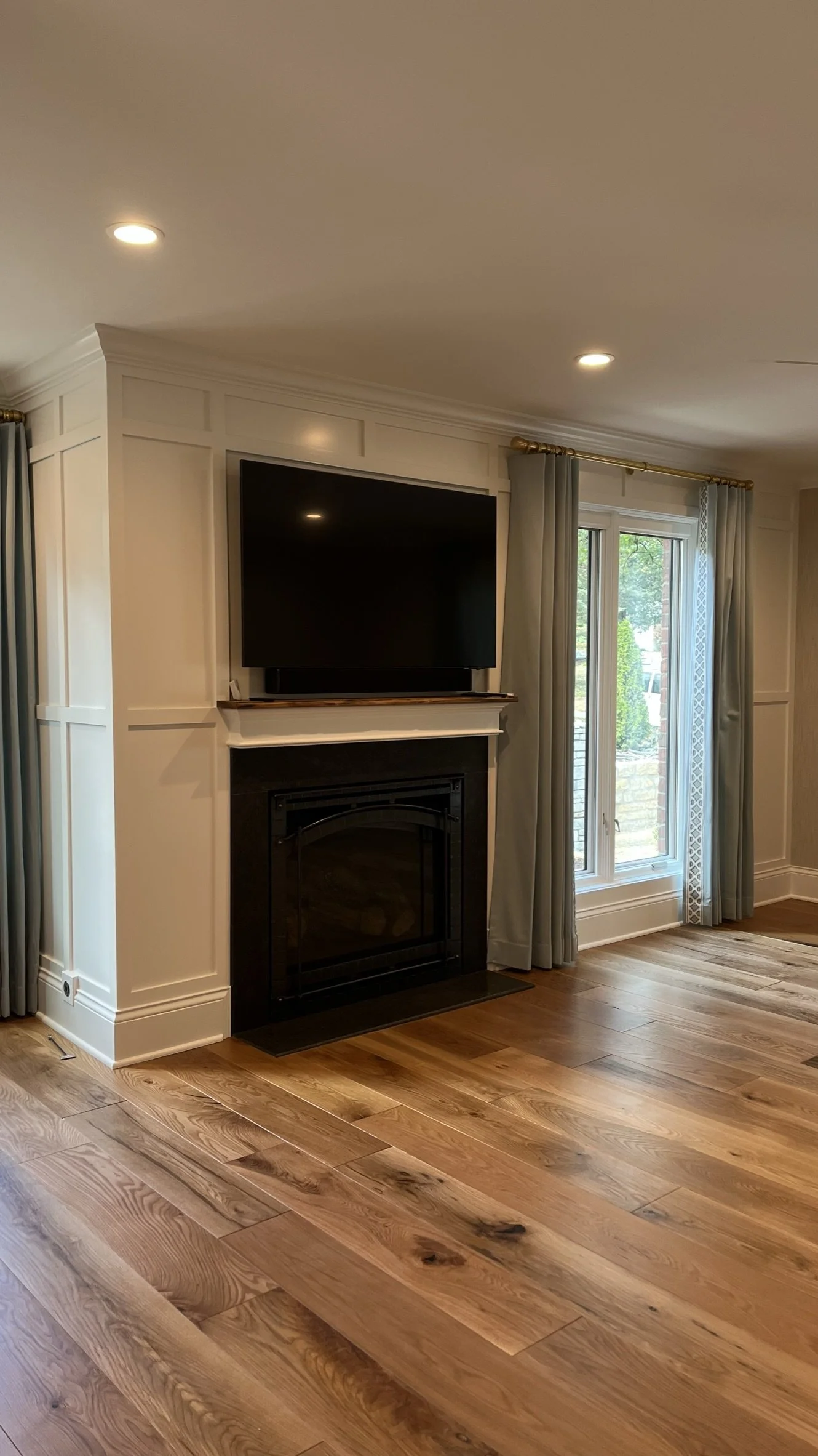 Living room with wood floor, white paneled walls, a fireplace with a TV mounted above, a window with blue curtains, and recessed ceiling lights.