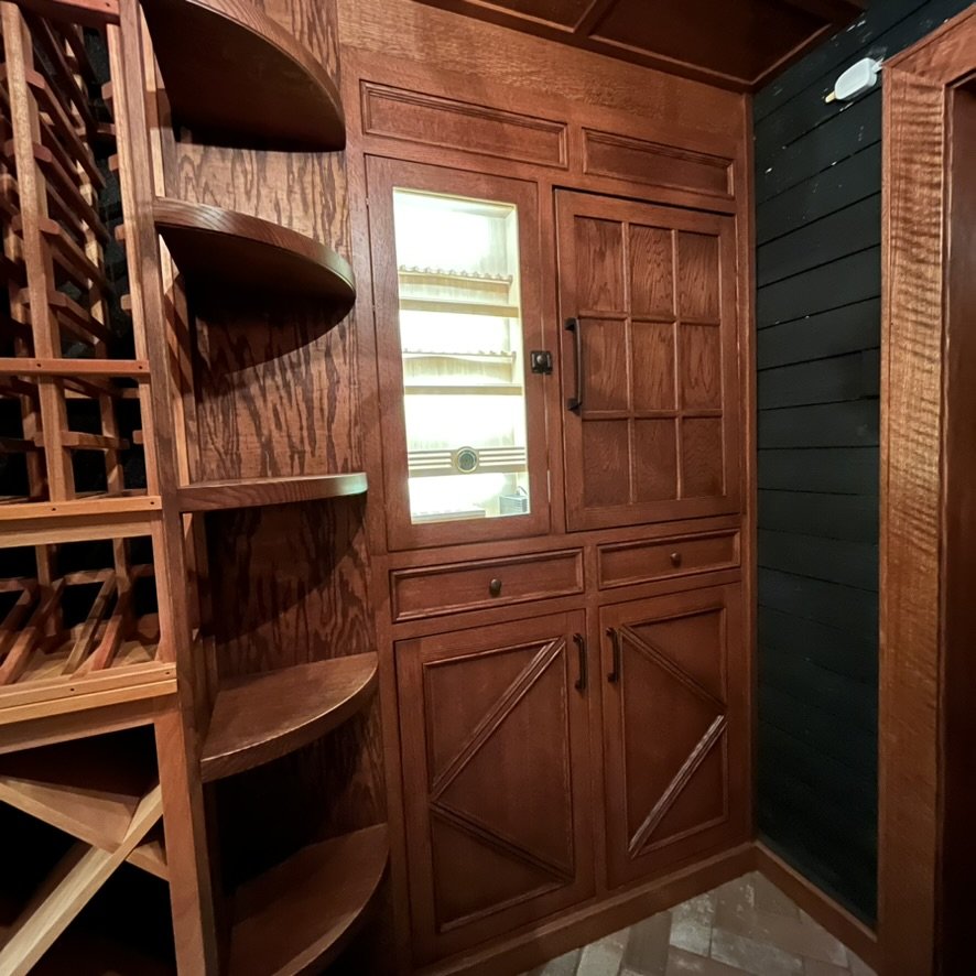 Wooden wine storage cabinet with glass-paneled door, drawers, and shelves adjacent to a wine rack in a cozy, wood-paneled room.