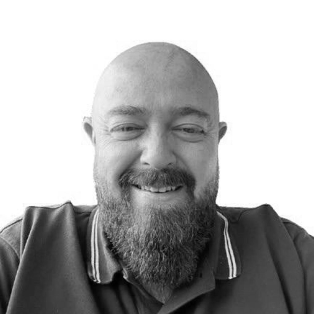 A smiling man with a beard and bald head, wearing a dark polo shirt with a white collar, against a plain background.